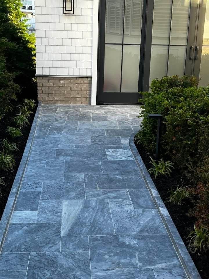 Blue stone pathway leading to a building entrance with dark framed glass doors, bordered by landscaping.