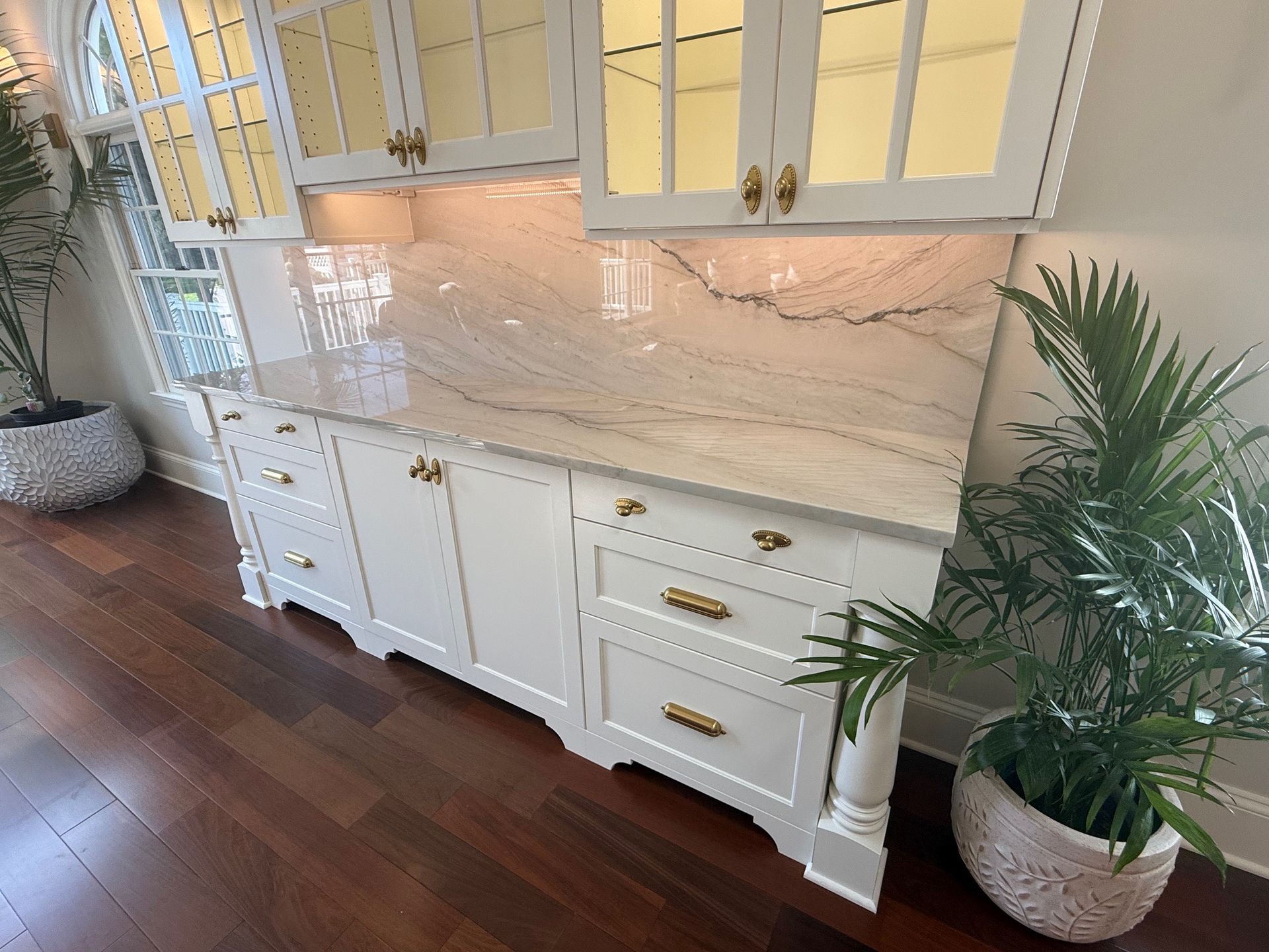 White cabinet with marble countertop, gold hardware, and plants, in a room with dark wood floor.