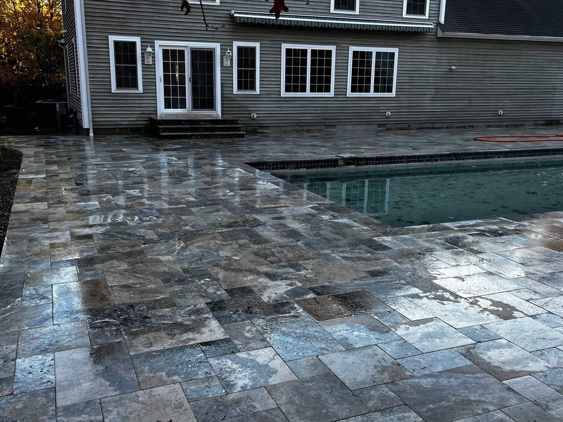 Patio with wet paving stones next to a pool and house, reflecting the sky.