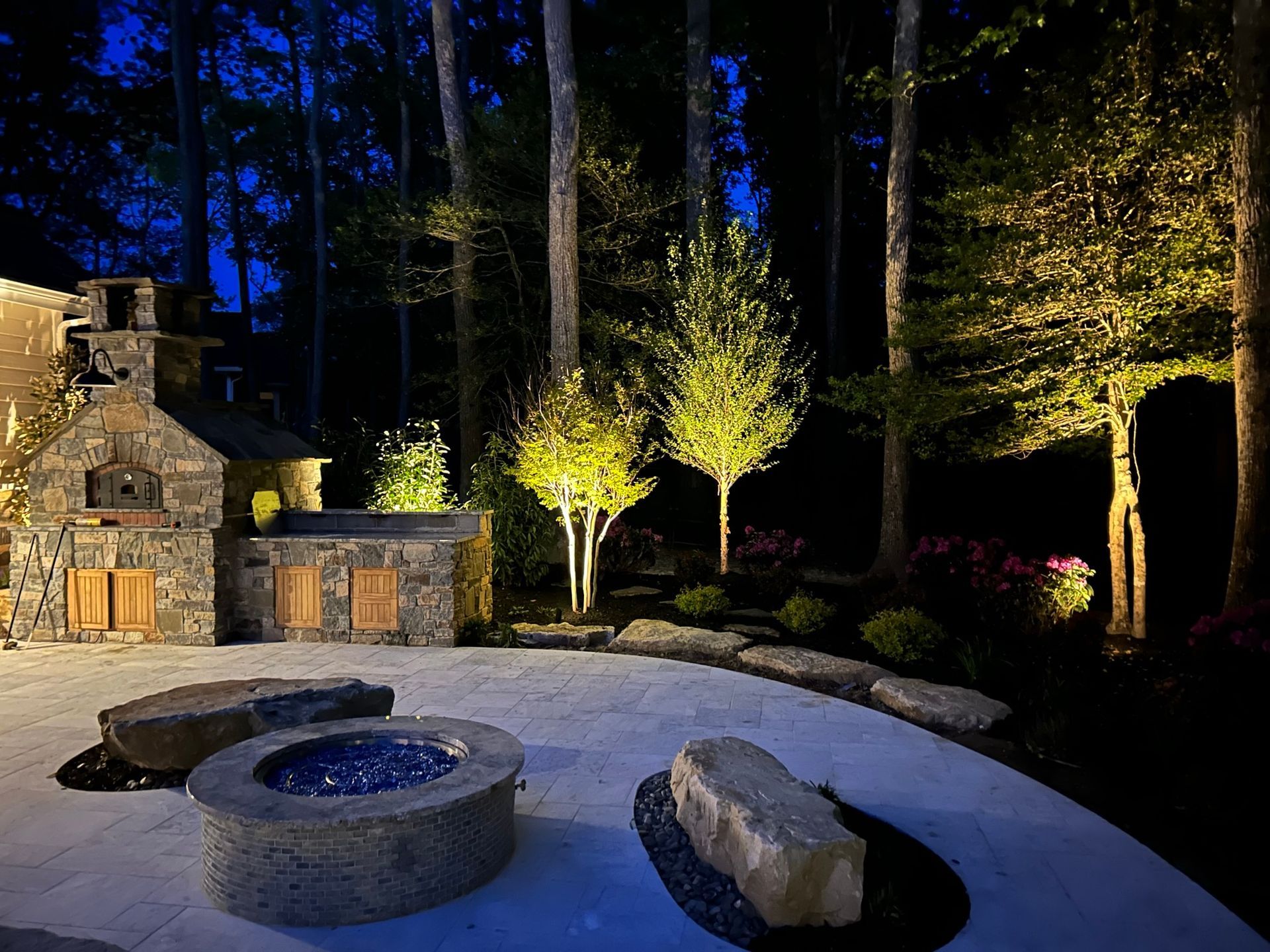 Nighttime backyard scene with illuminated stone patio, outdoor kitchen, fire pit, trees, and garden beds.