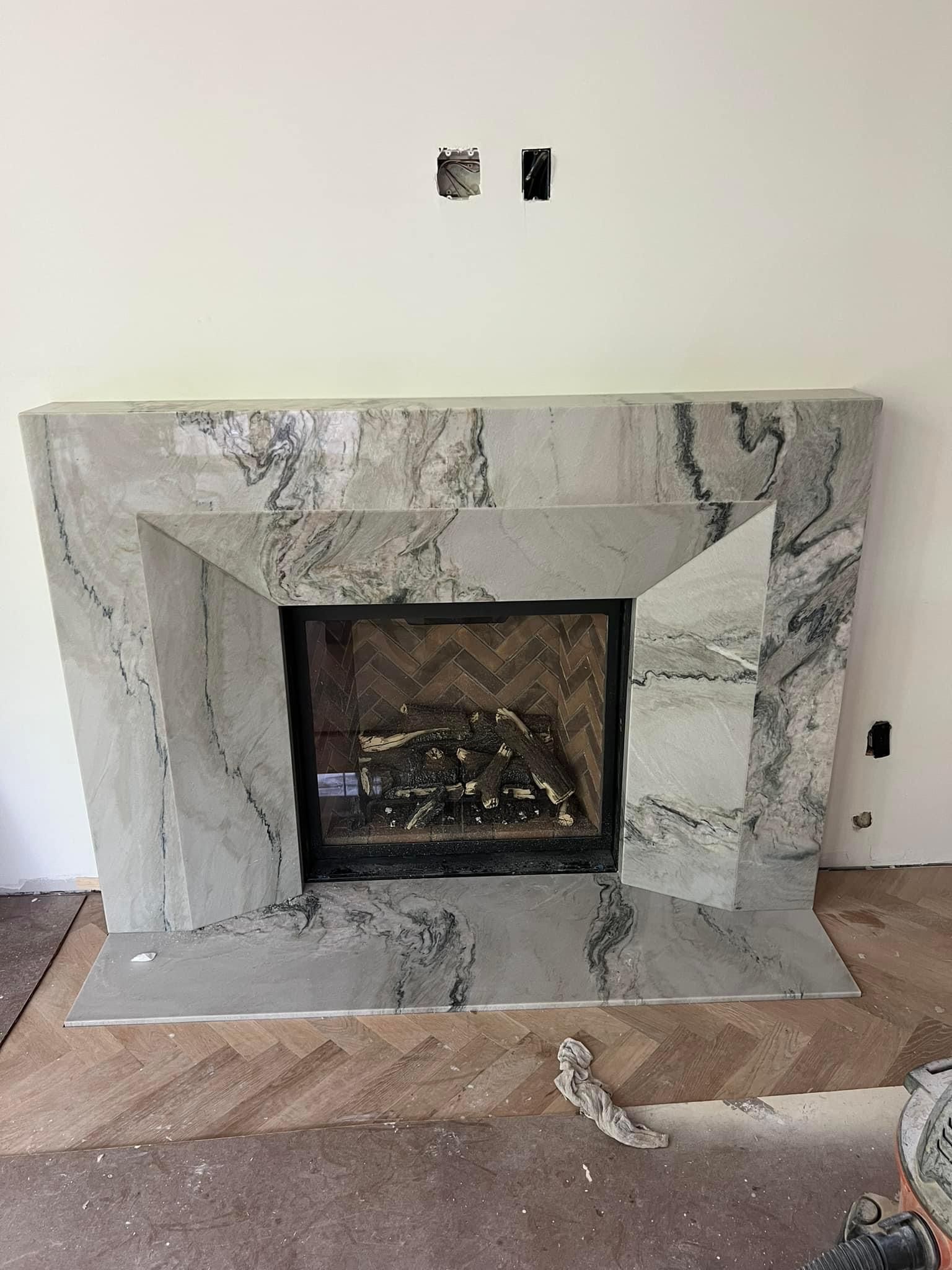 Stone fireplace with grey marble, on a herringbone floor, and an empty wall above it.