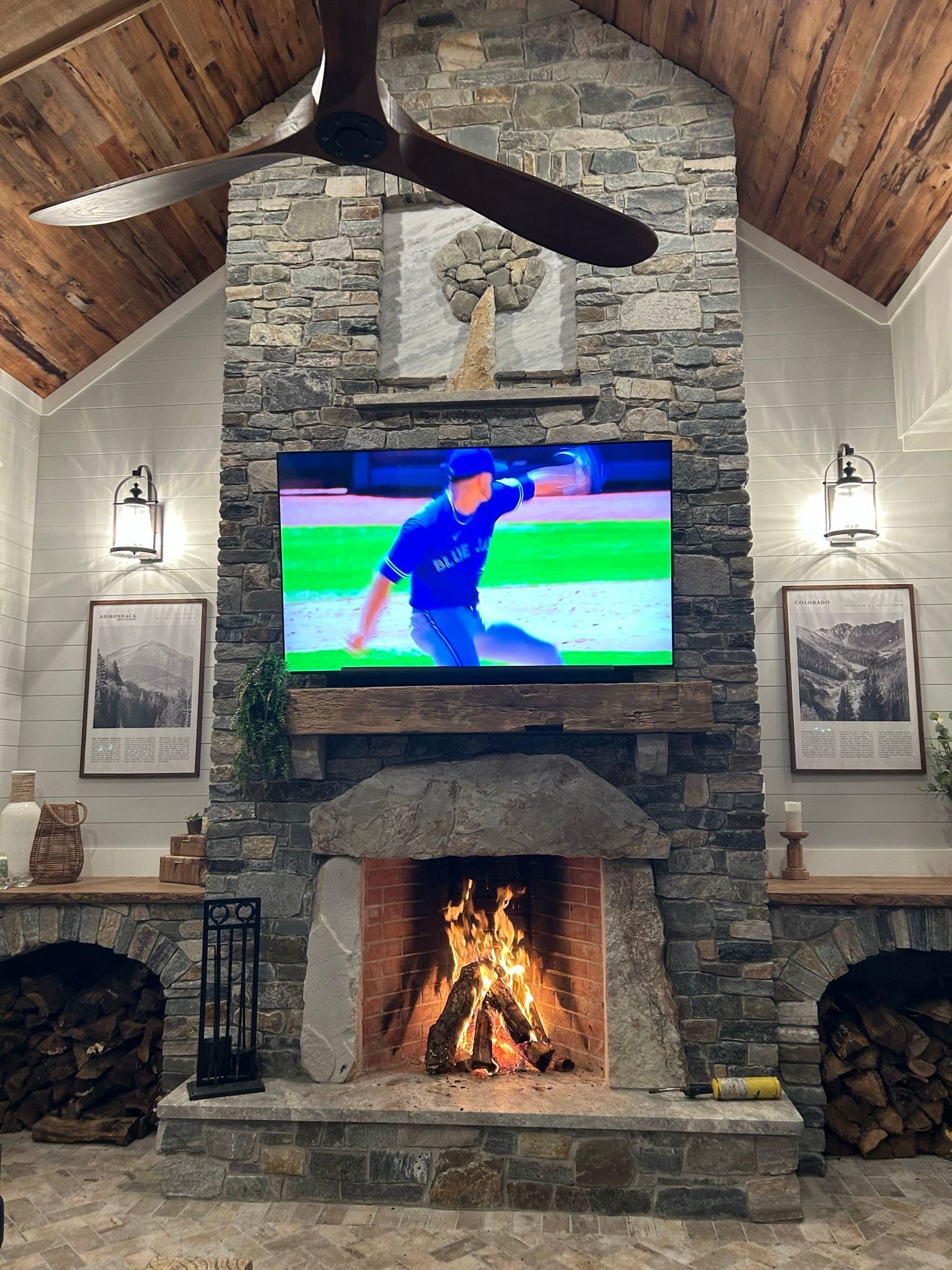 Fireplace with lit fire, TV showing baseball player, wood-paneled ceiling, stone walls, sconces, framed art, and wood storage.