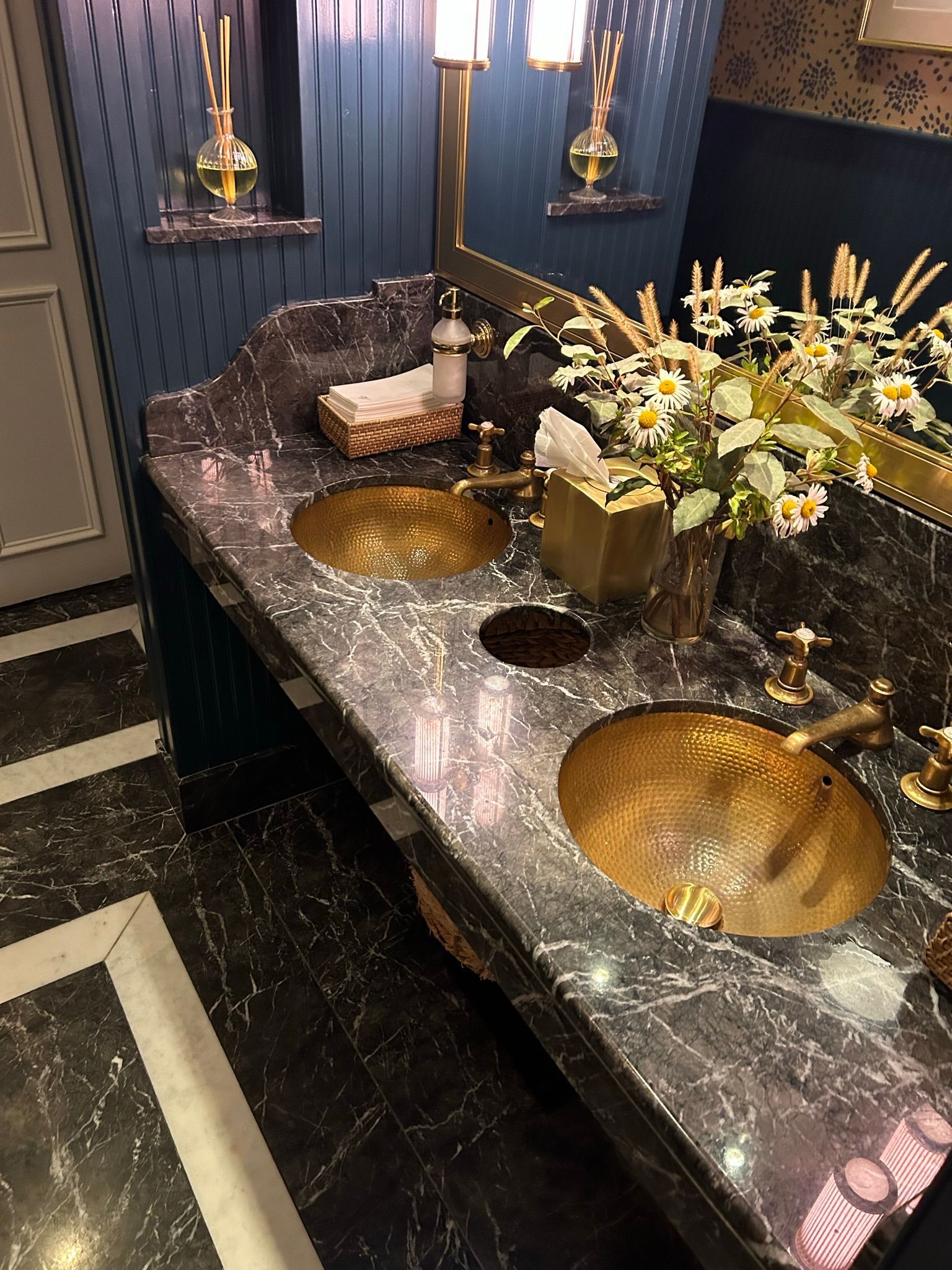 Elegant bathroom with marble counter, gold sinks, flowers, gold fixtures, blue walls.