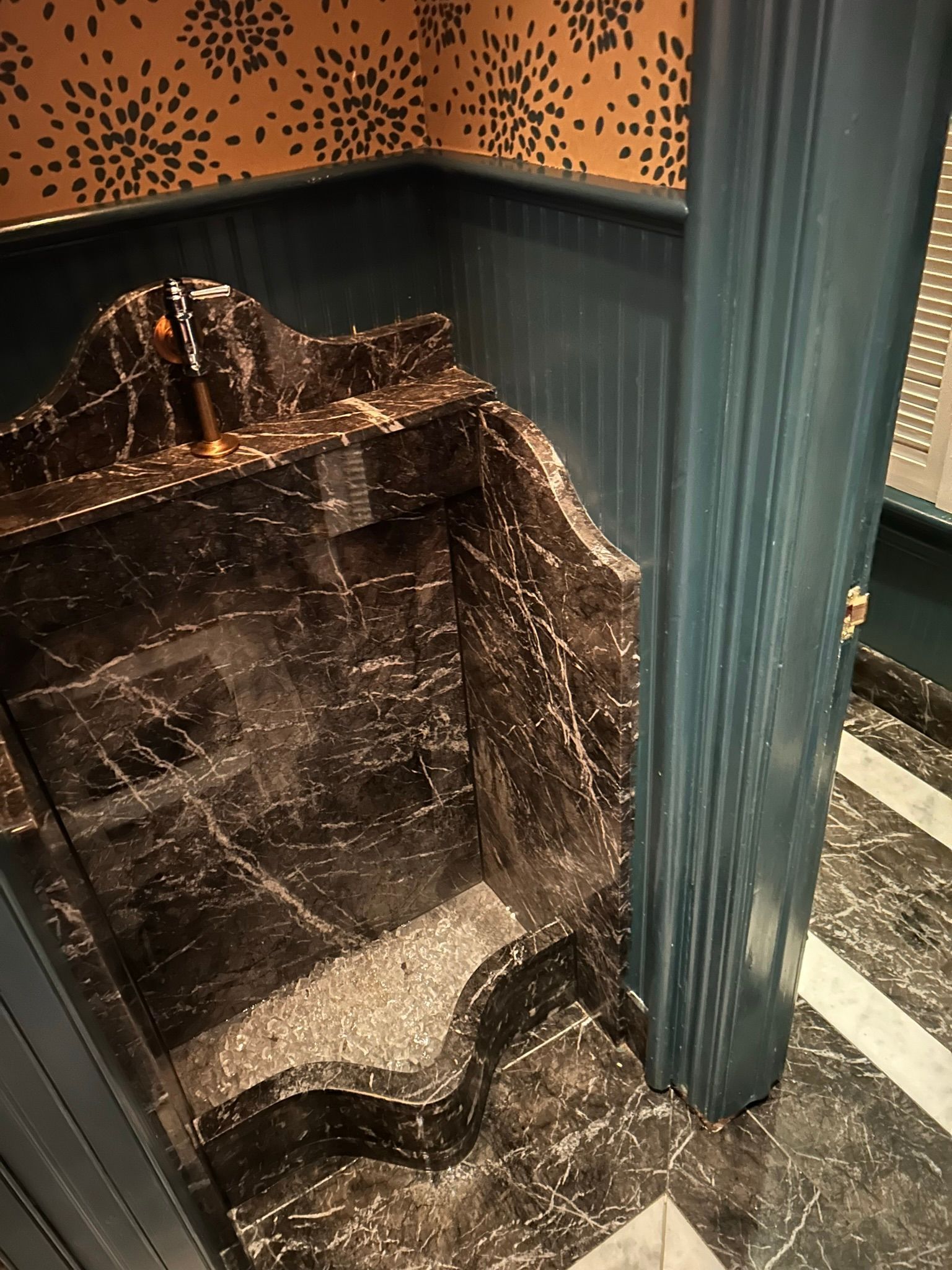 Dark marble corner sink with decorative scalloped edges in a room with green and patterned wallpaper.