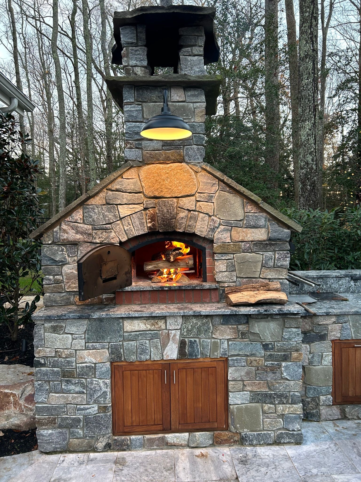 Stone outdoor pizza oven with burning fire, wooden doors, and pendant light.