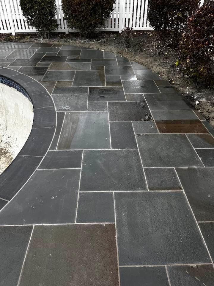 Dark stone patio with curved edge by a pool, leading to a white picket fence, bushes on either side.