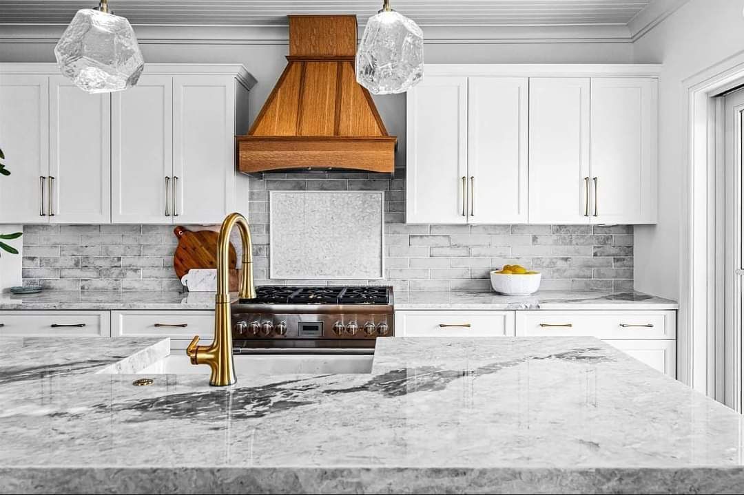 White kitchen with marble countertops, gold faucet, and wooden range hood.