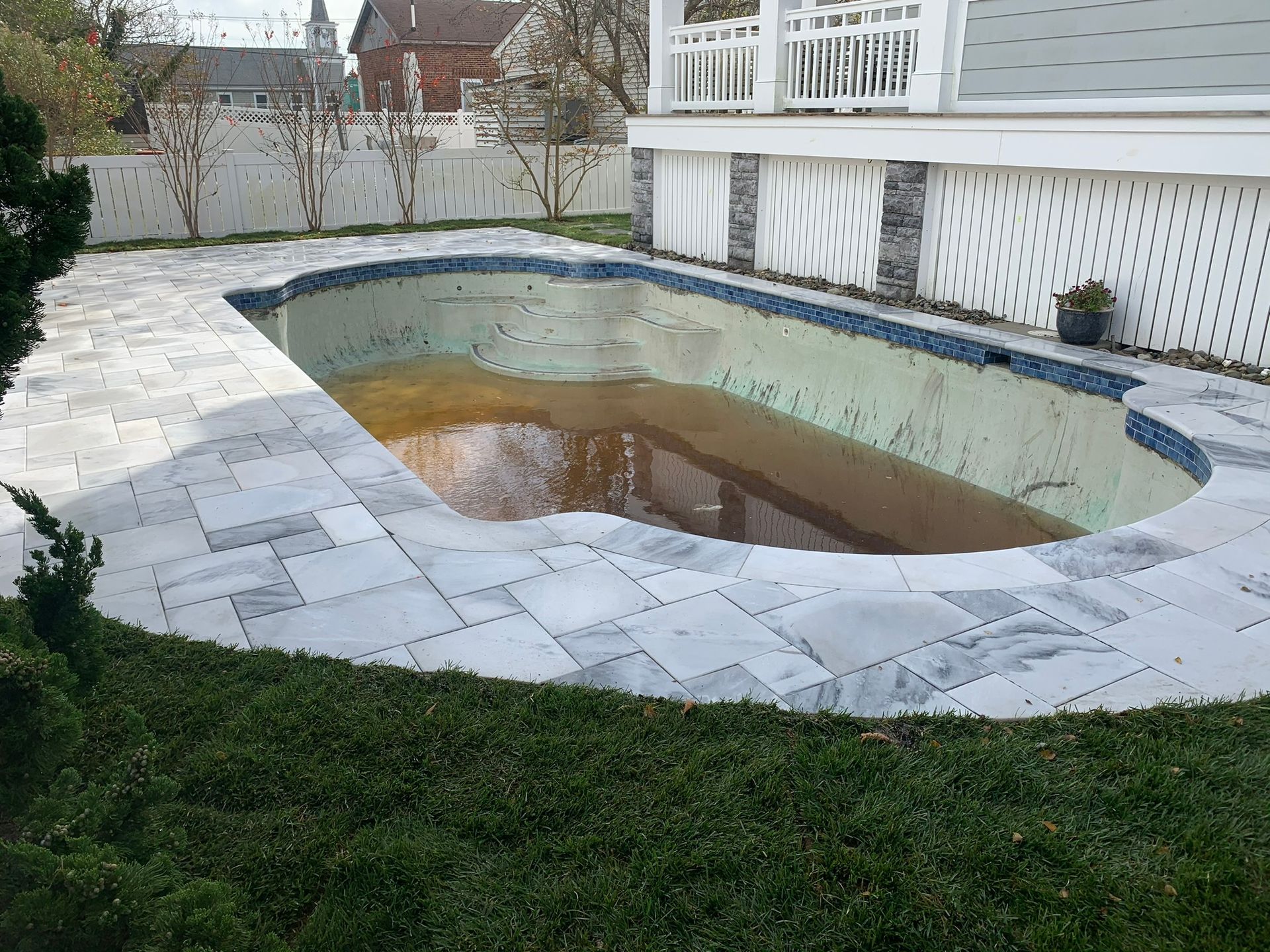 Empty, dirty swimming pool with blue-tiled edges and surrounding gray stone patio; green grass and white picket fence.