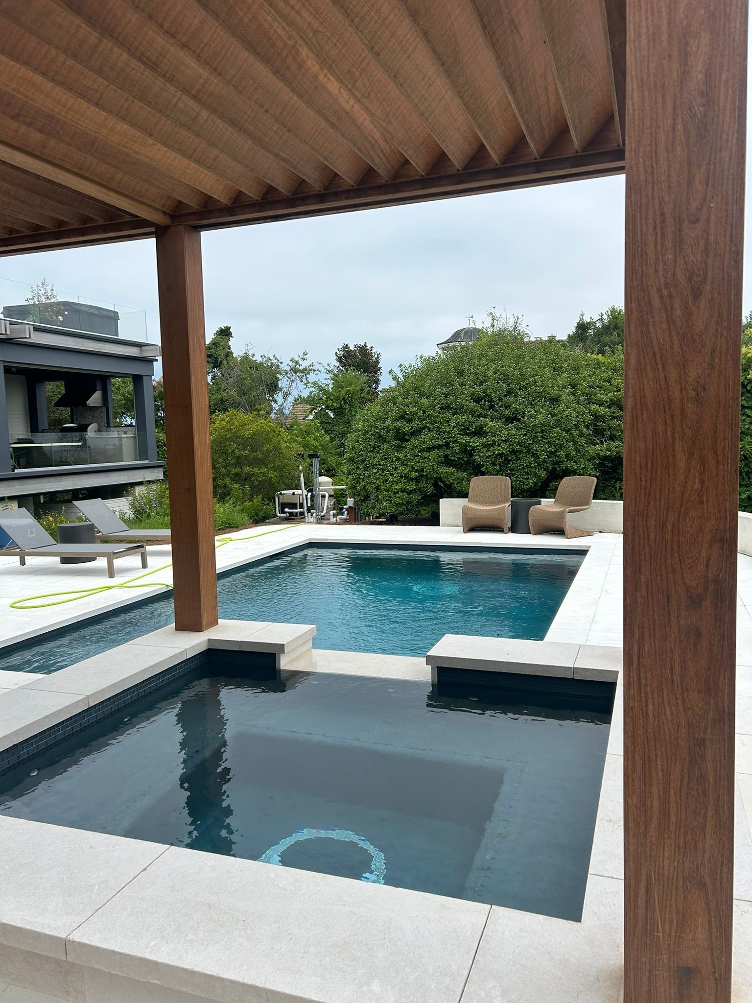 Pool and spa under a wooden pergola, overlooking greenery and another building.