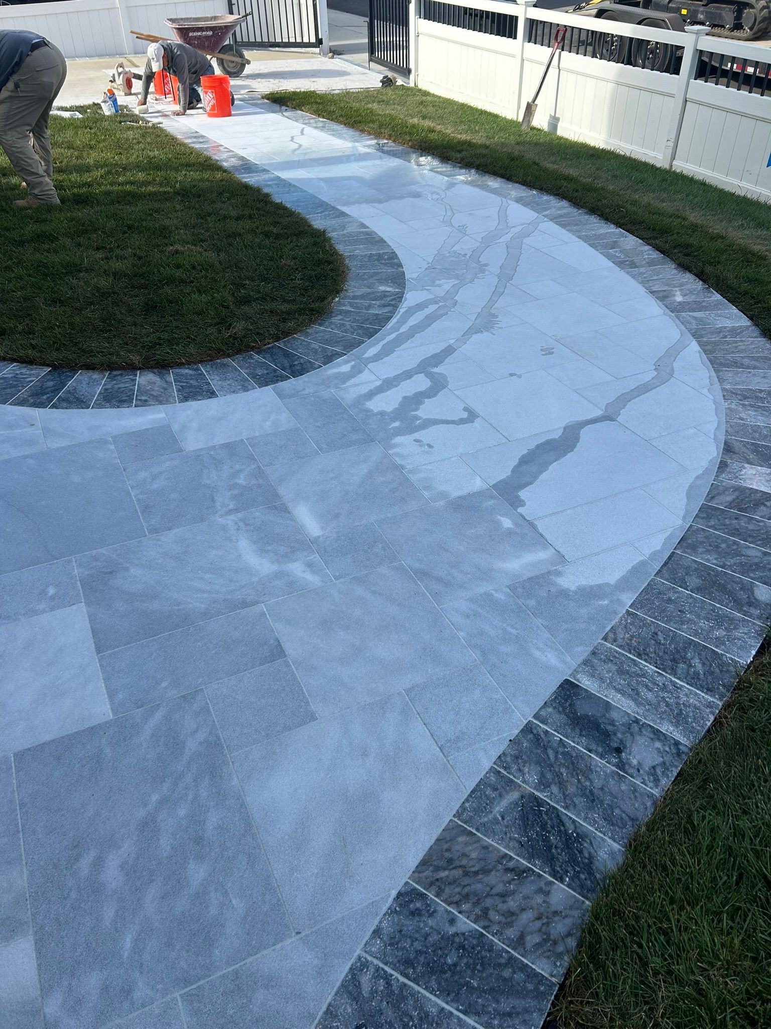 Curving stone pathway bordered by dark bricks and grass, with a person working in the background.