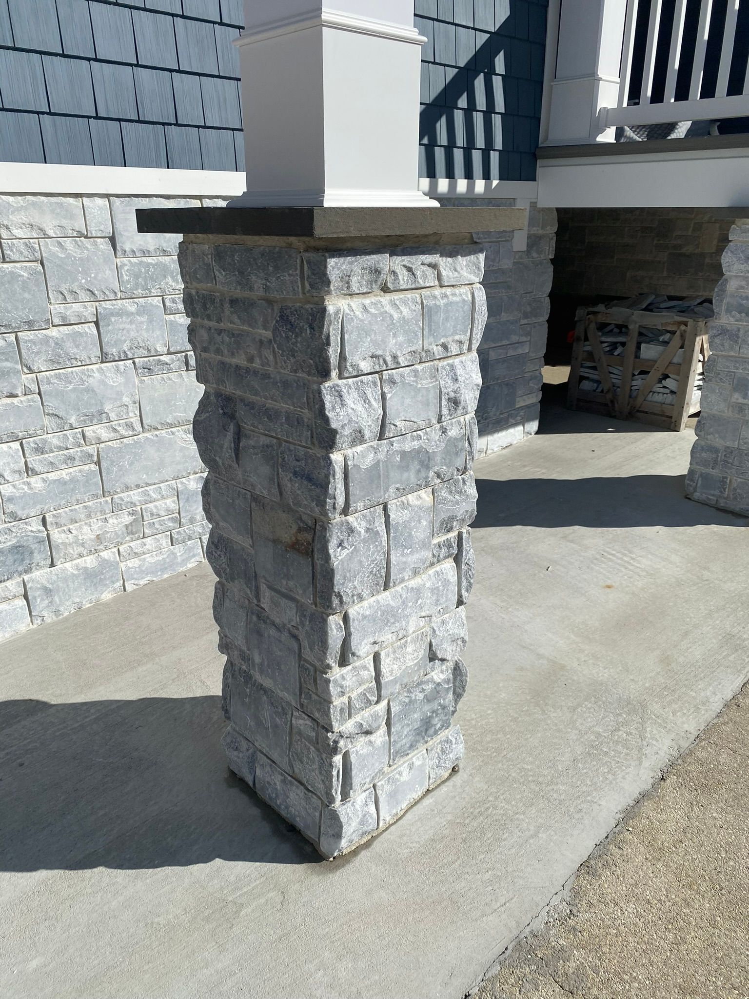 Gray stone pillar supporting a white beam, set on a concrete porch, with a blue-sided house in the background.