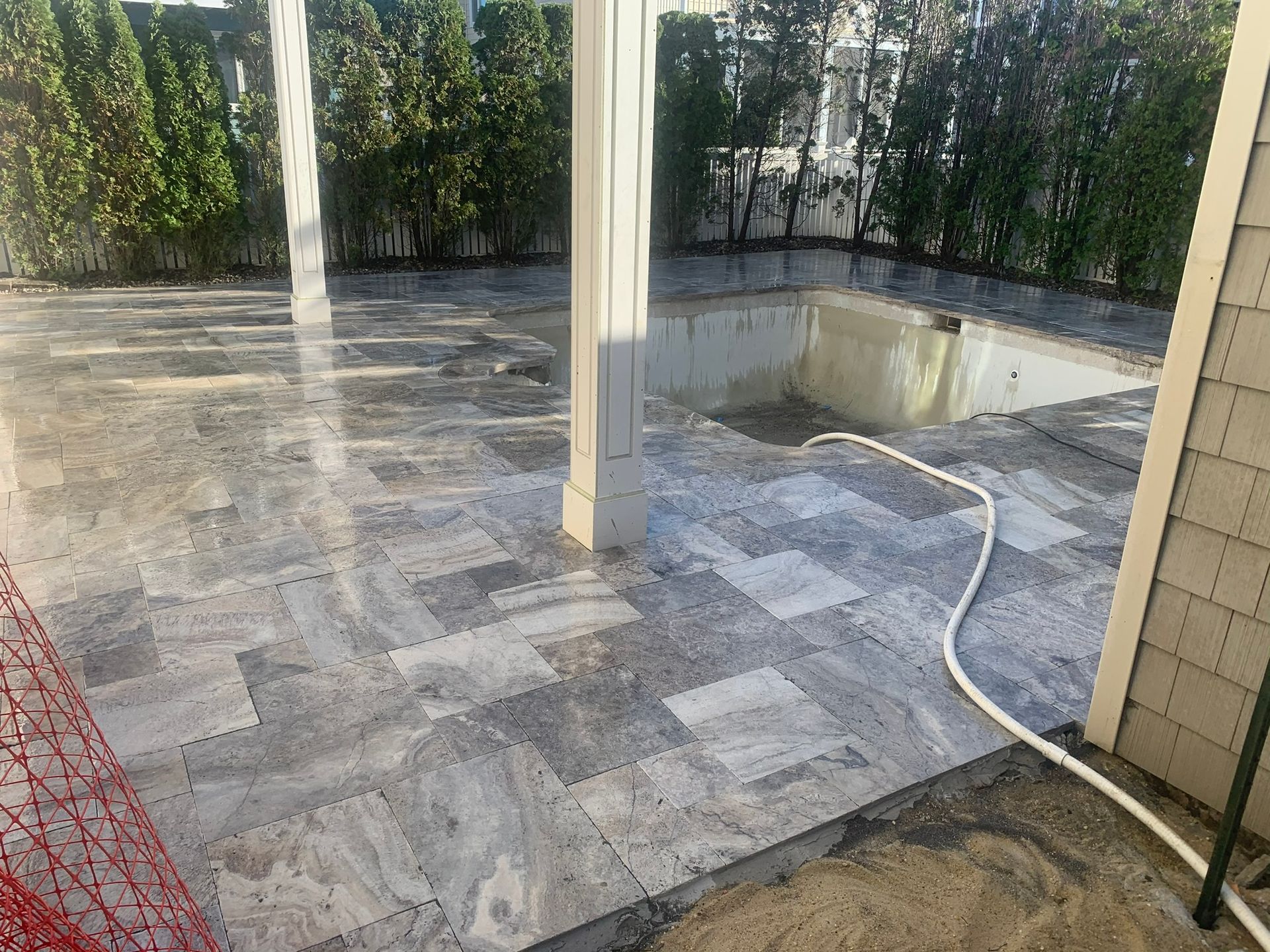 Grey stone patio being constructed with a concrete pool area and green hedges in the background.