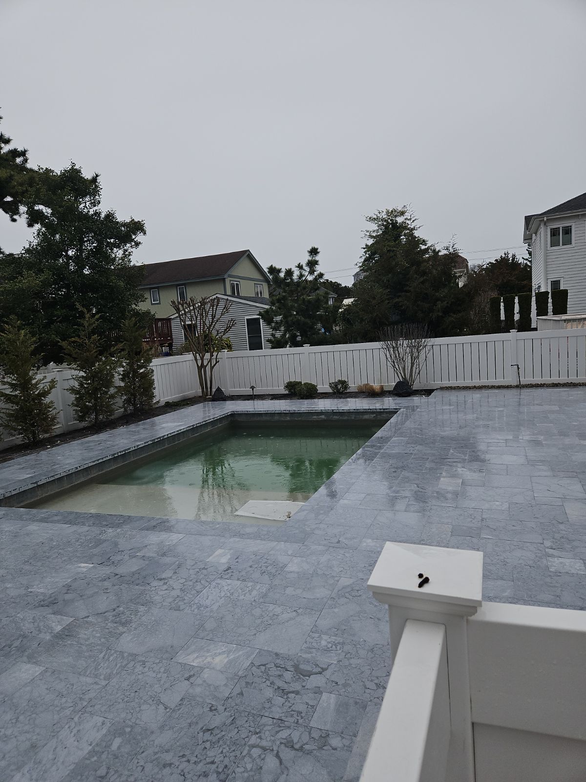 A gray, rectangular pool with murky green water sits in a backyard, surrounded by a stone patio. White fence in the background.