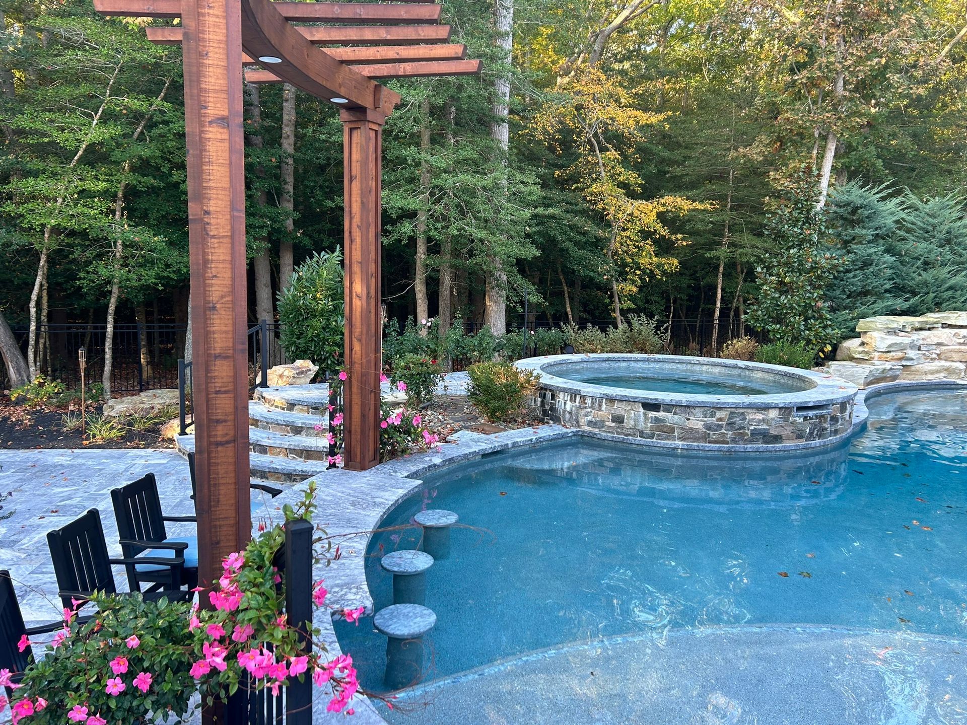 Pool with a hot tub, pergola, and bar stools, surrounded by trees.