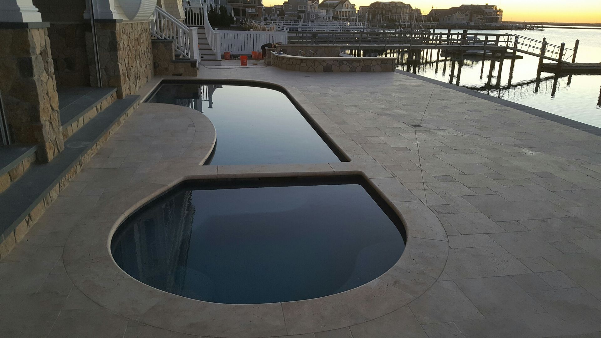A pool with a curved edge, next to a dock on the water, and stone steps.