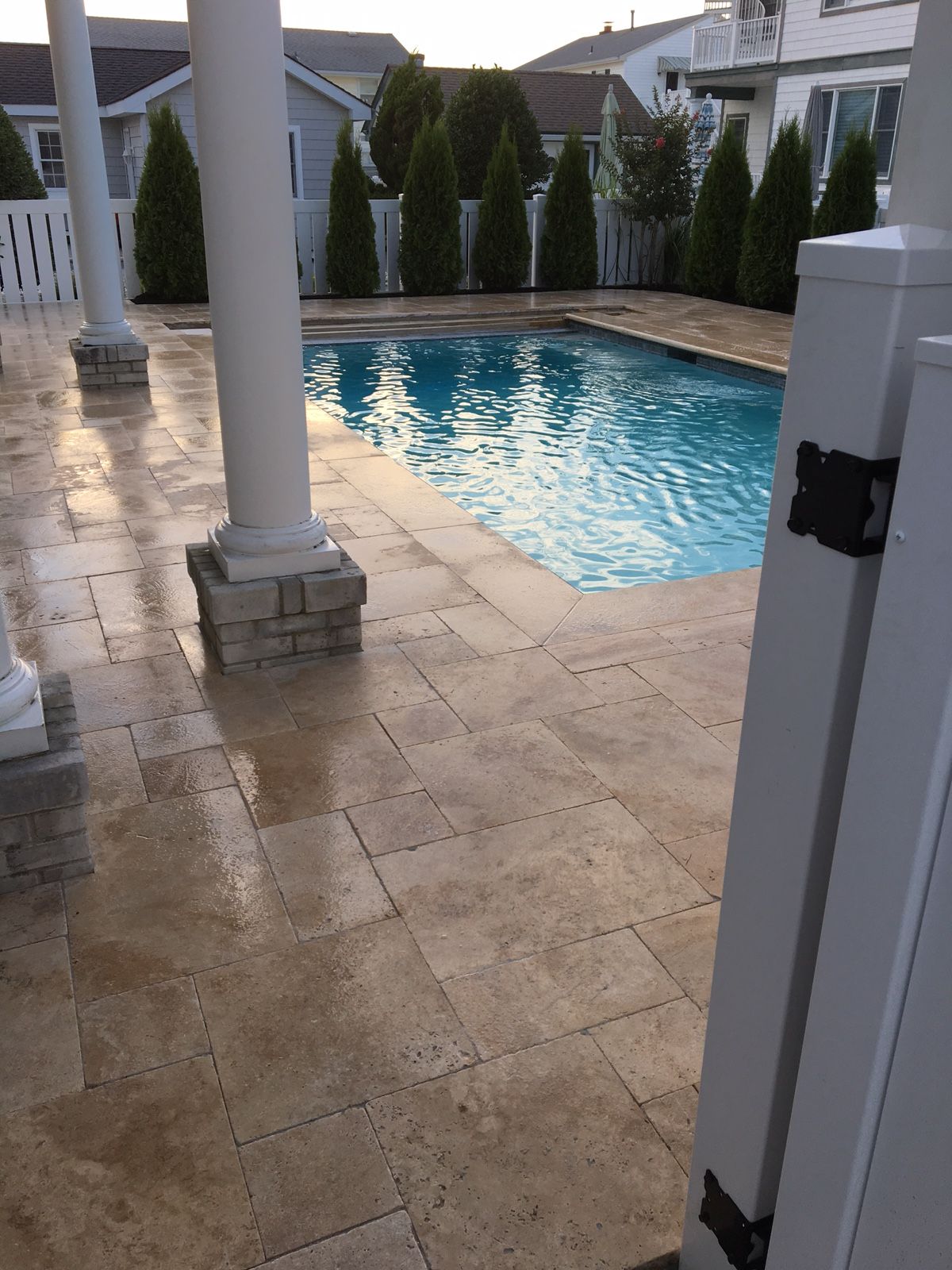 Poolside scene with travertine pavers, white columns, and a blue pool. Green trees line the fence in the background.