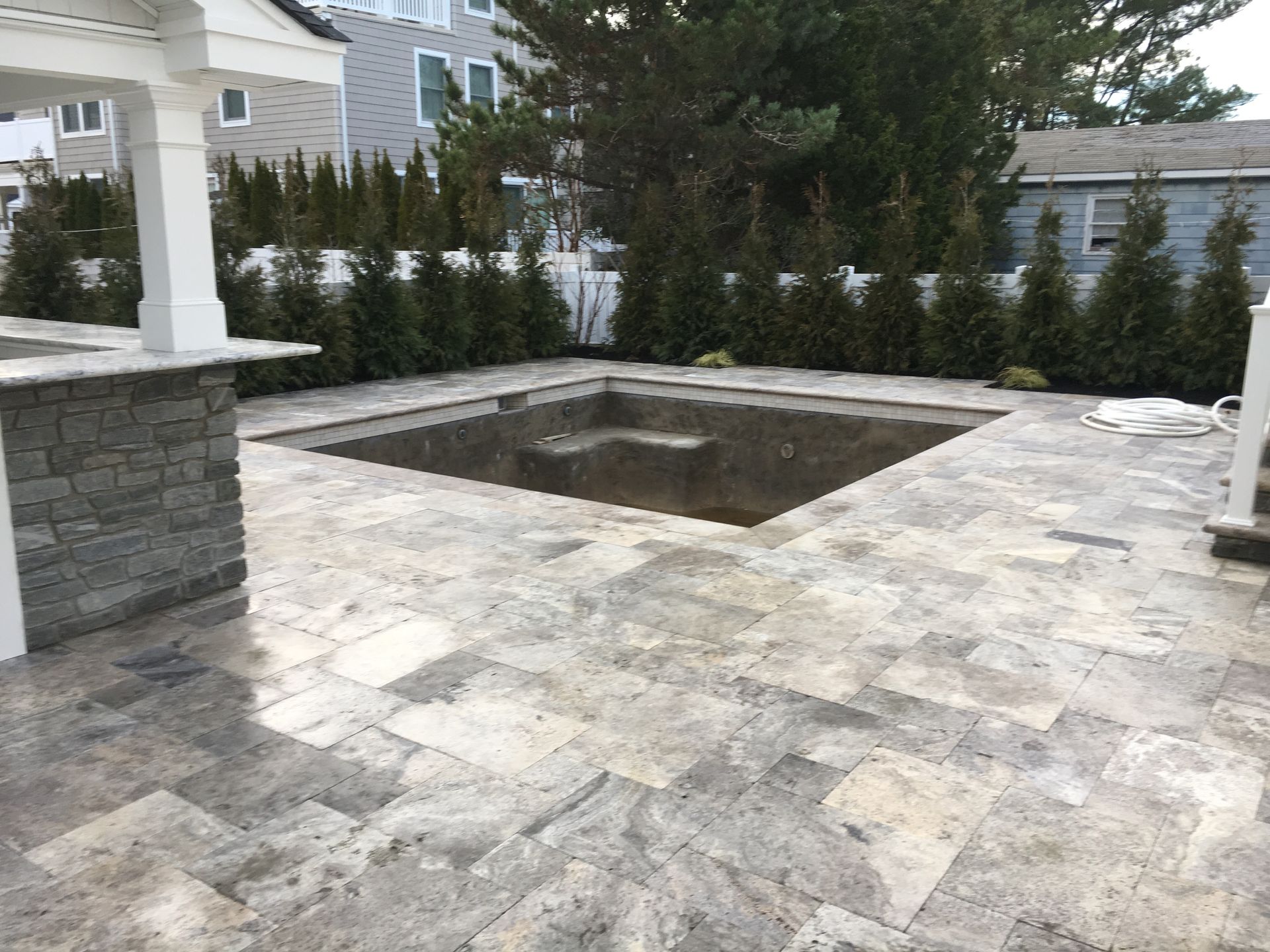 A square, empty pool surrounded by gray stone patio. Green hedges and a white structure are in the background.