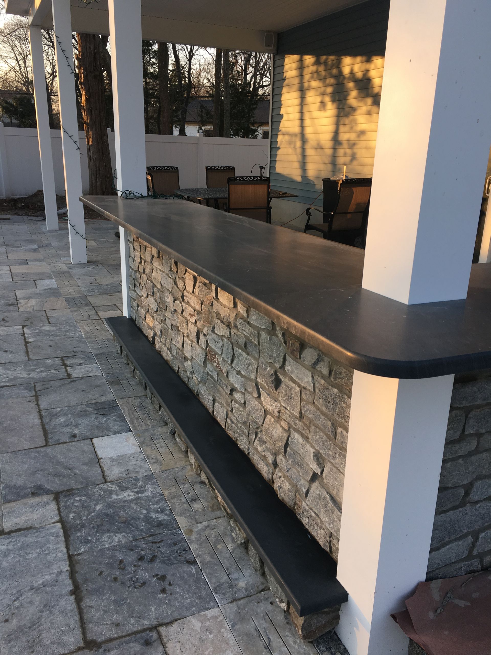 Outdoor bar with stone facade, gray countertop, and white pillars, set on a stone patio.