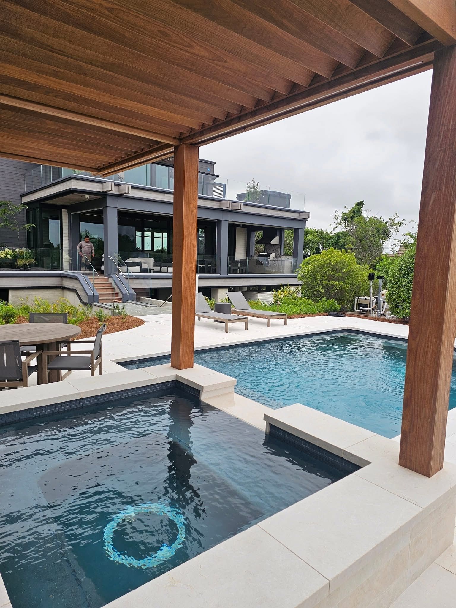 Pool area with a house in the background; wooden pergola, blue pool, dark gray house, cloudy sky.
