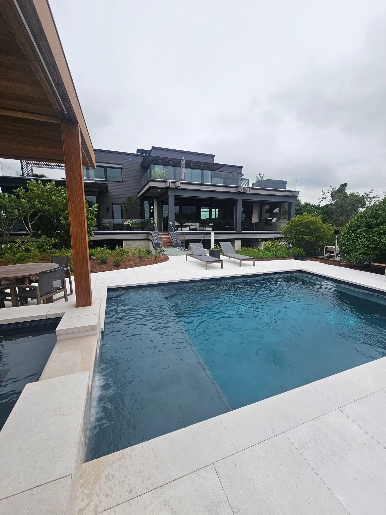 Luxurious home with a pool and lounge chairs, surrounded by lush greenery under cloudy skies.