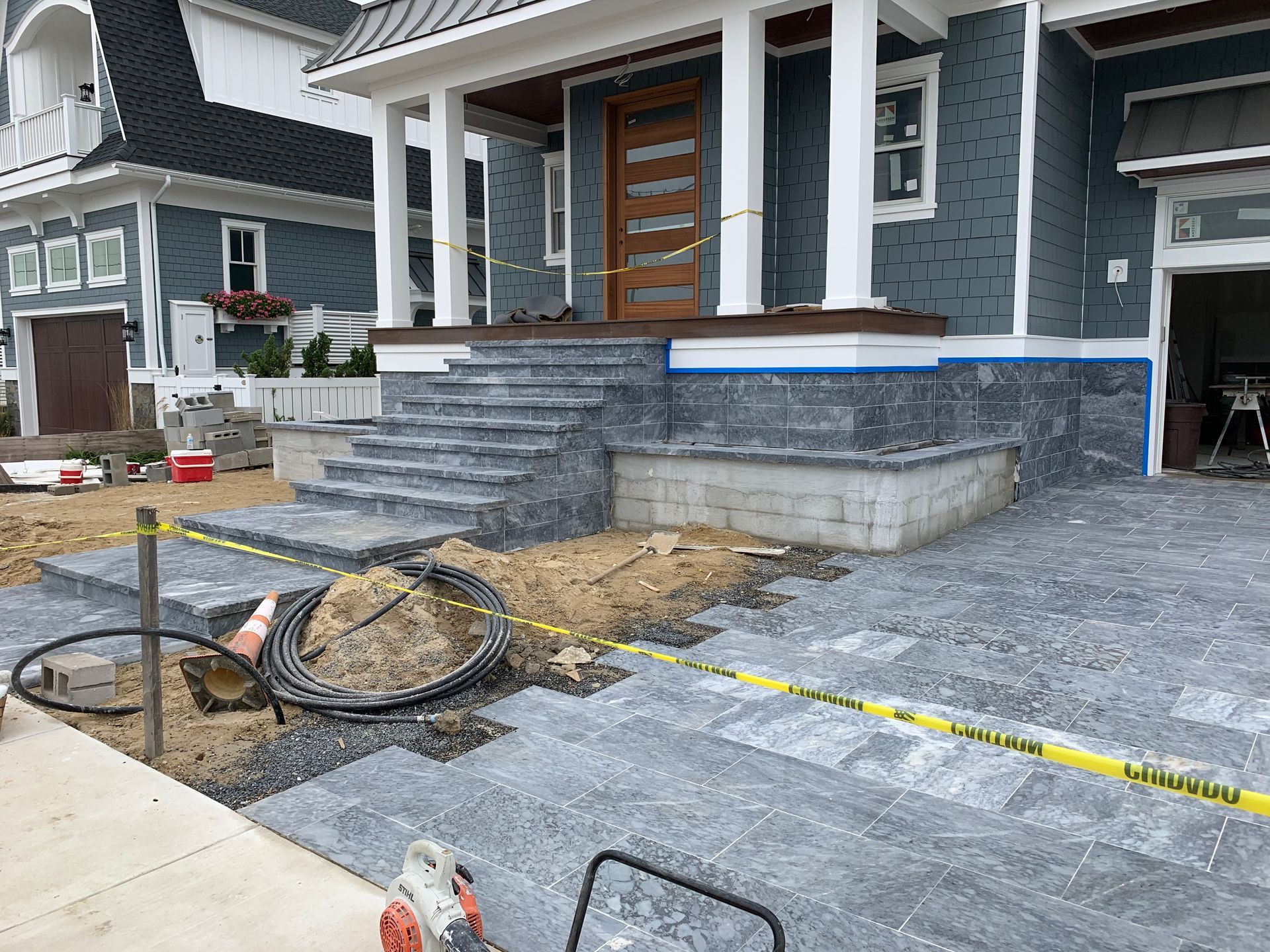 Construction site with gray pavers, steps, and blue house siding. Yellow caution tape.
