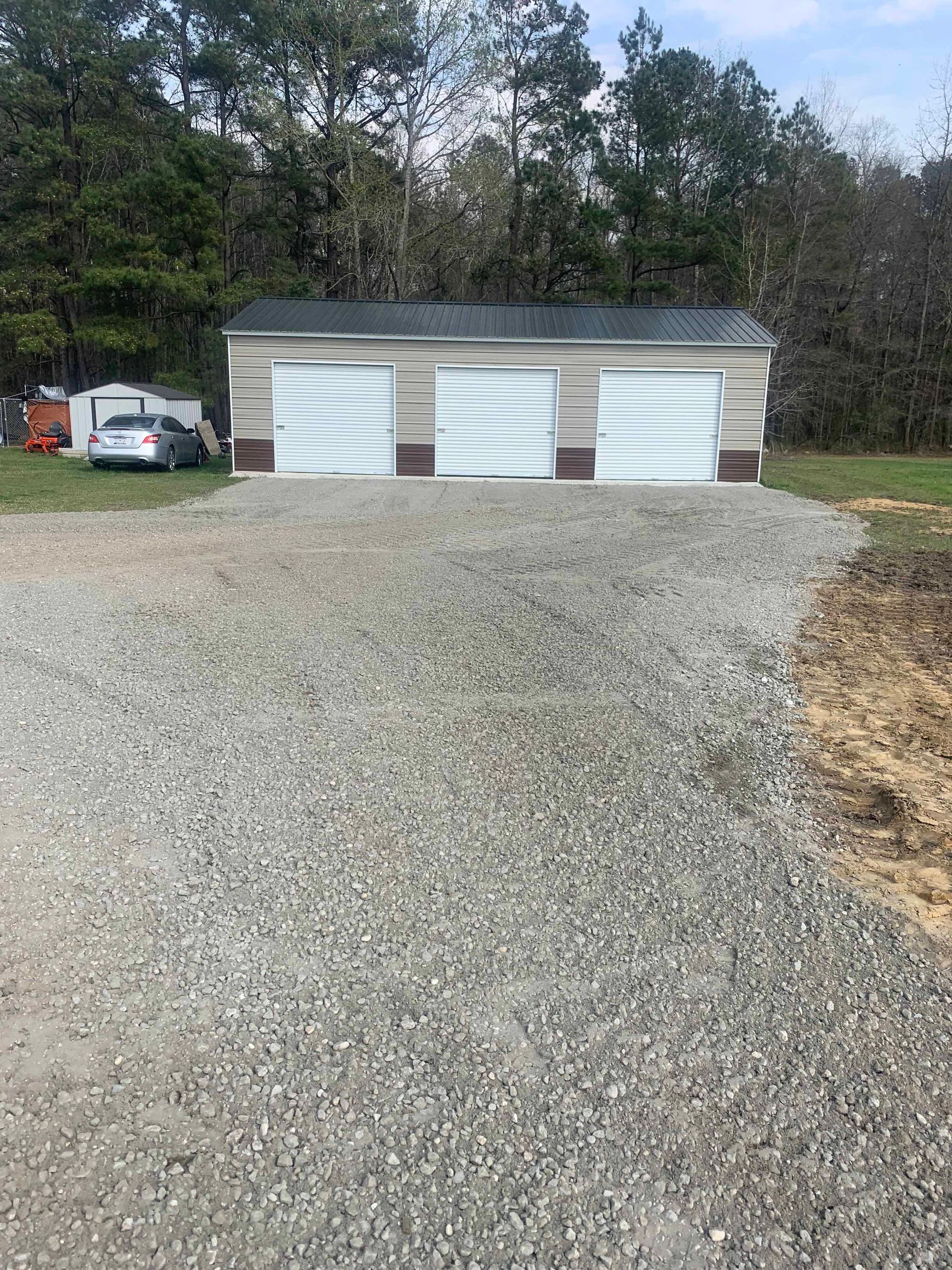 Gravel driveway leading to a three-bay garage with white doors. A car and small building are visible in the distance.