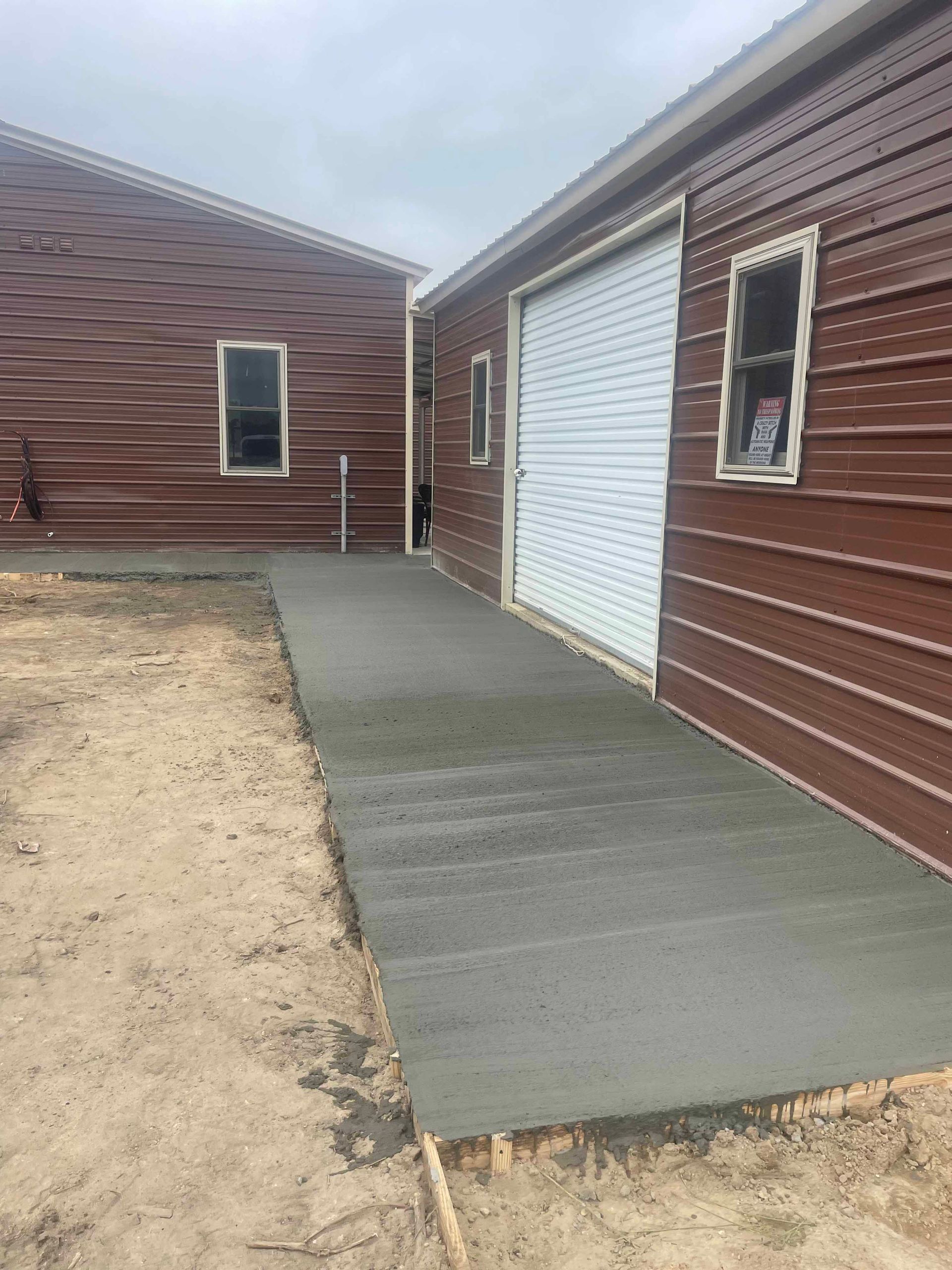 A newly poured concrete walkway between two brown-sided buildings. The walkway is light gray, and the ground is dirt.