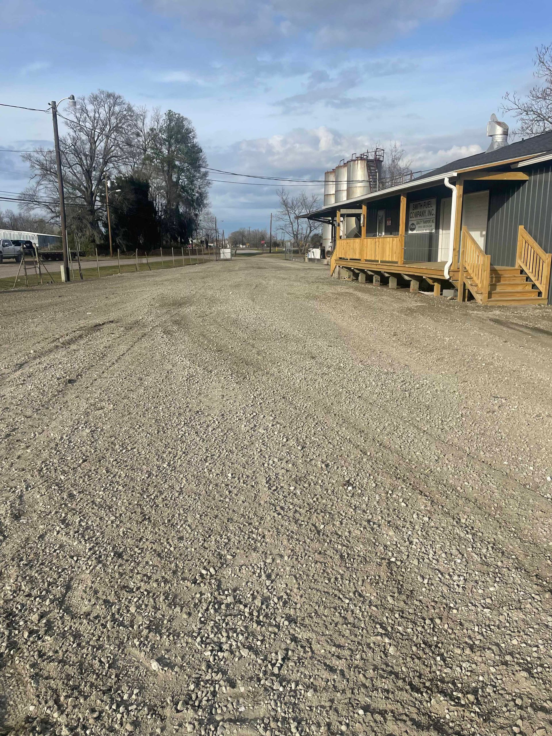 Gravel parking lot with a wooden porch building on the right. Bare trees and overcast sky in the background.
