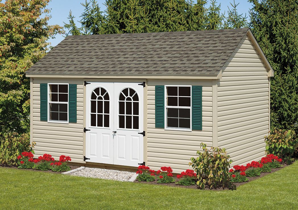 a white shed with green shutters is sitting in the middle of a lush green yard .