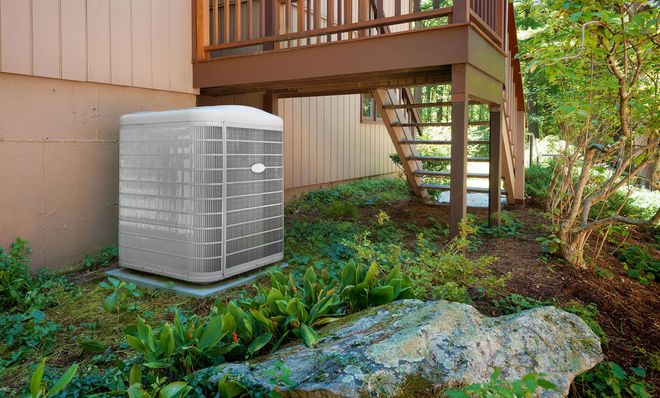 Air conditioner unit near a wooden deck and house, surrounded by greenery.