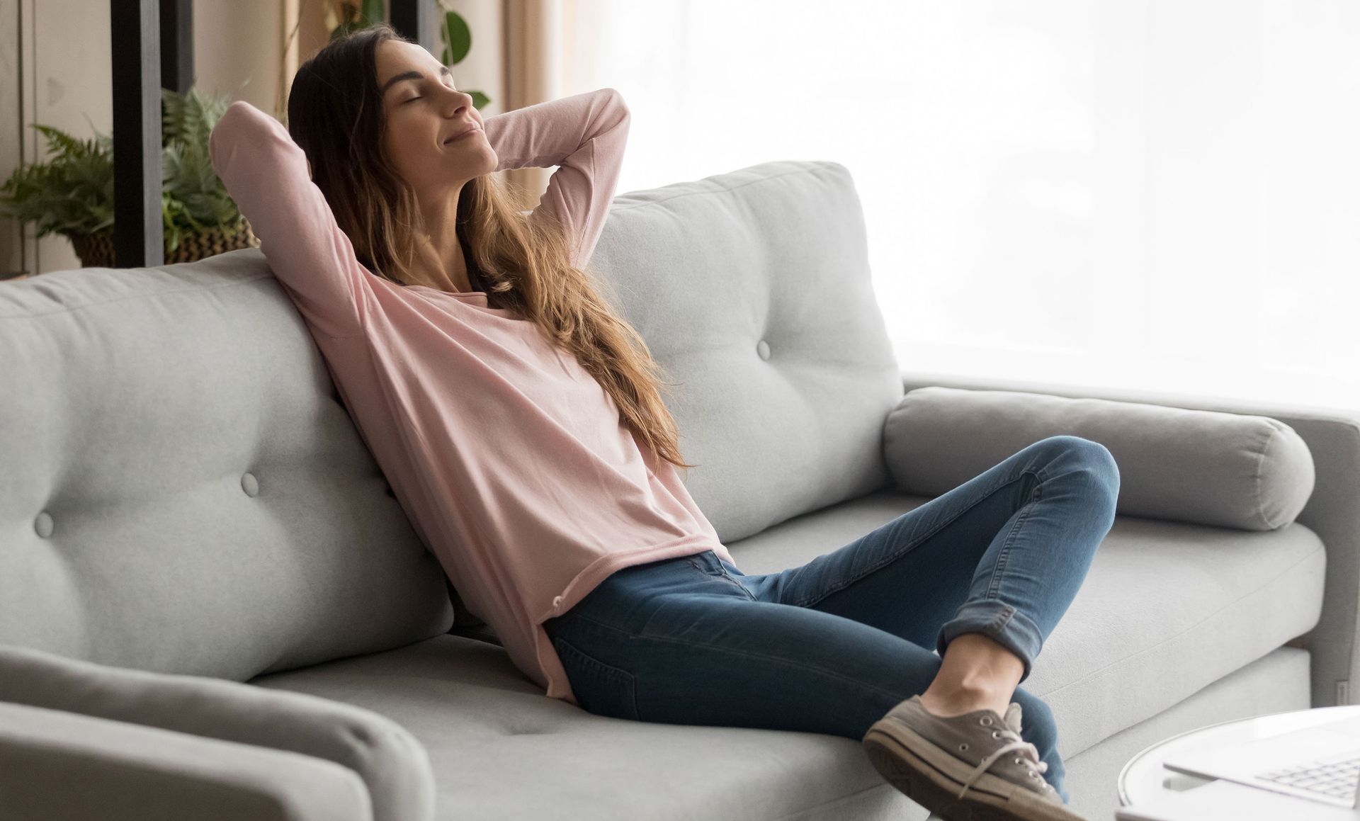 Woman relaxing on a grey sofa, arms behind head, eyes closed. Natural light, indoor setting.