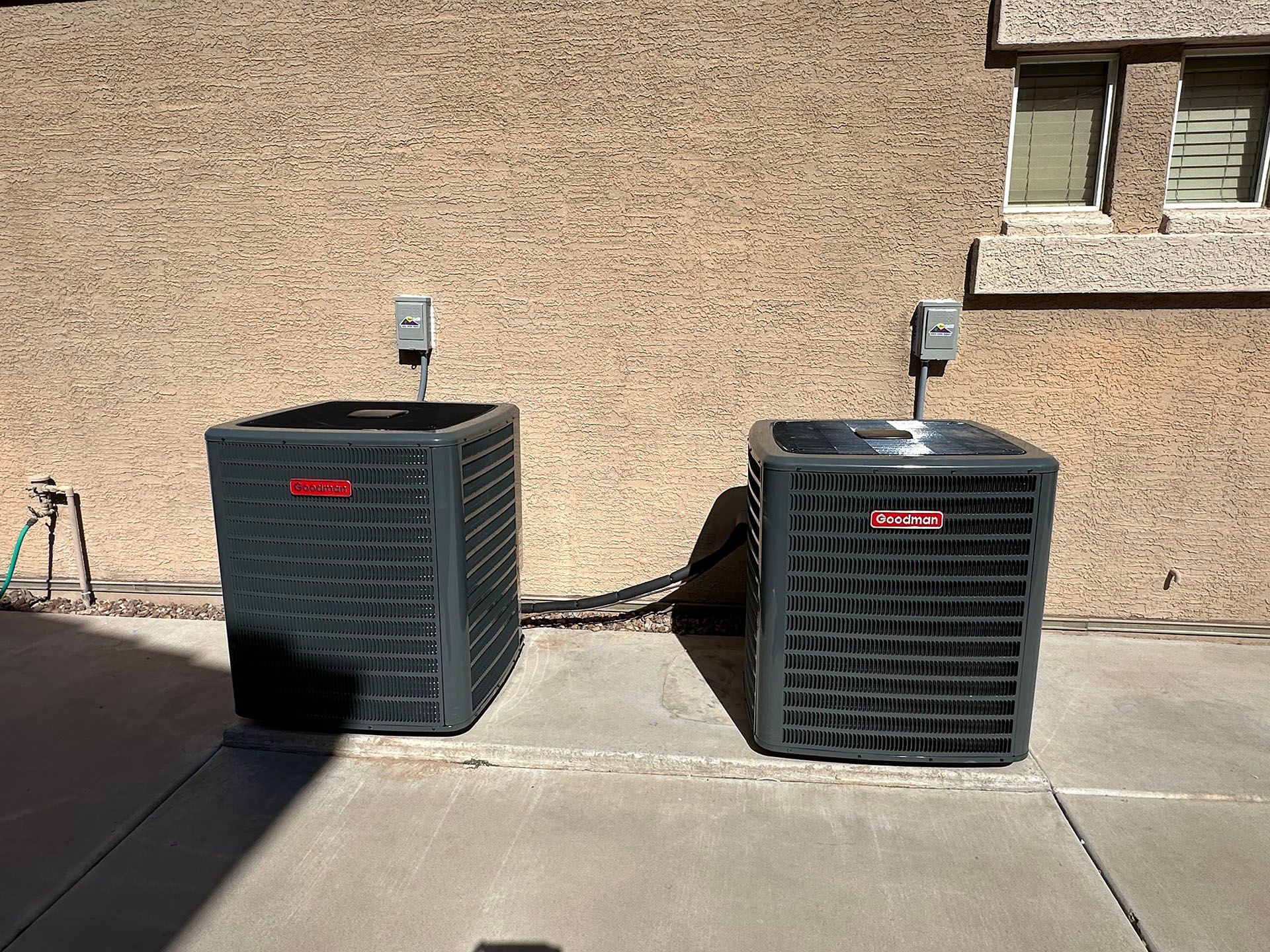 Two outdoor air conditioning units beside a tan stucco wall on a concrete patio.