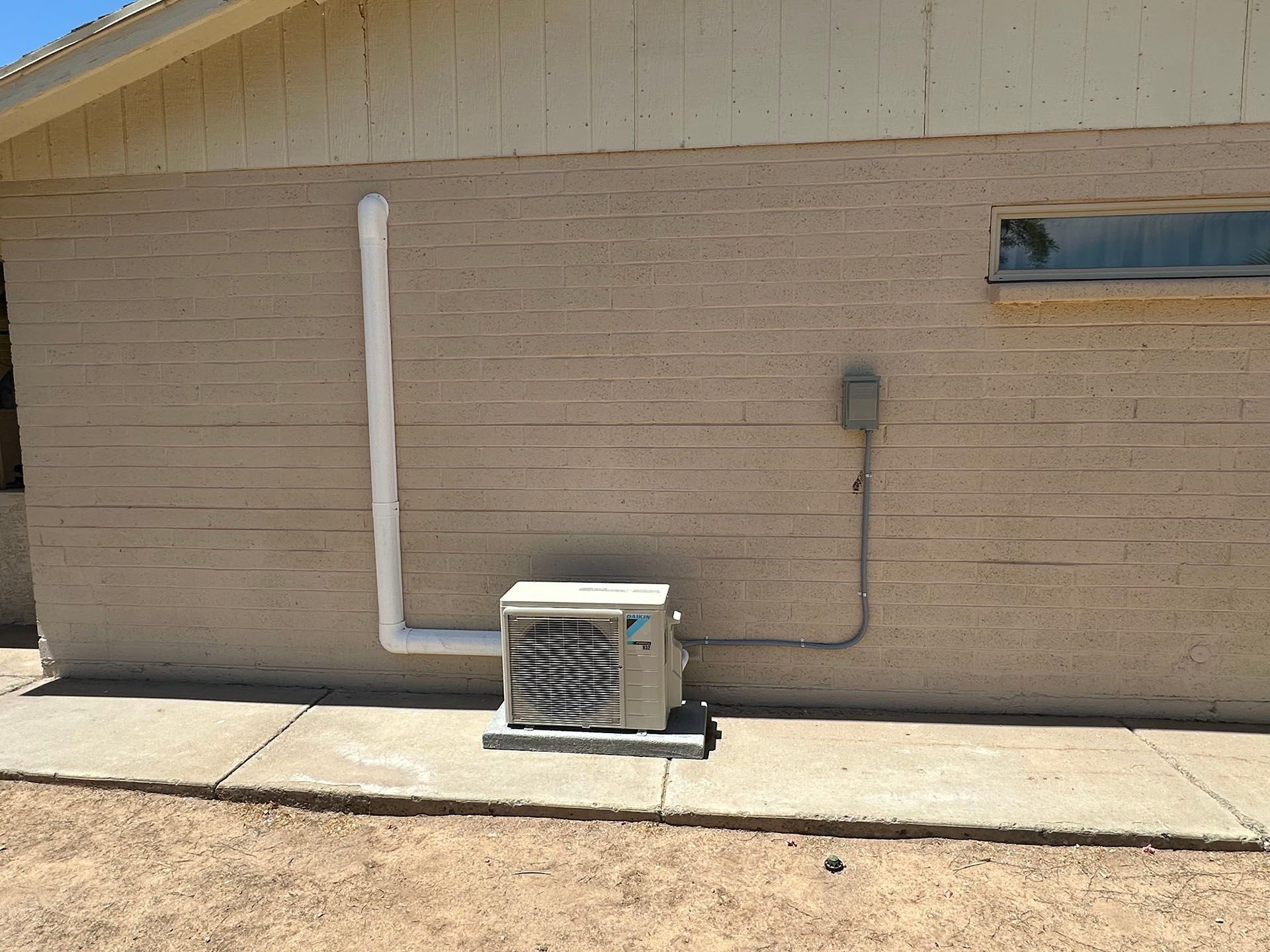 Brick building wall with a small outdoor AC unit, white conduit pipe, and narrow window above.