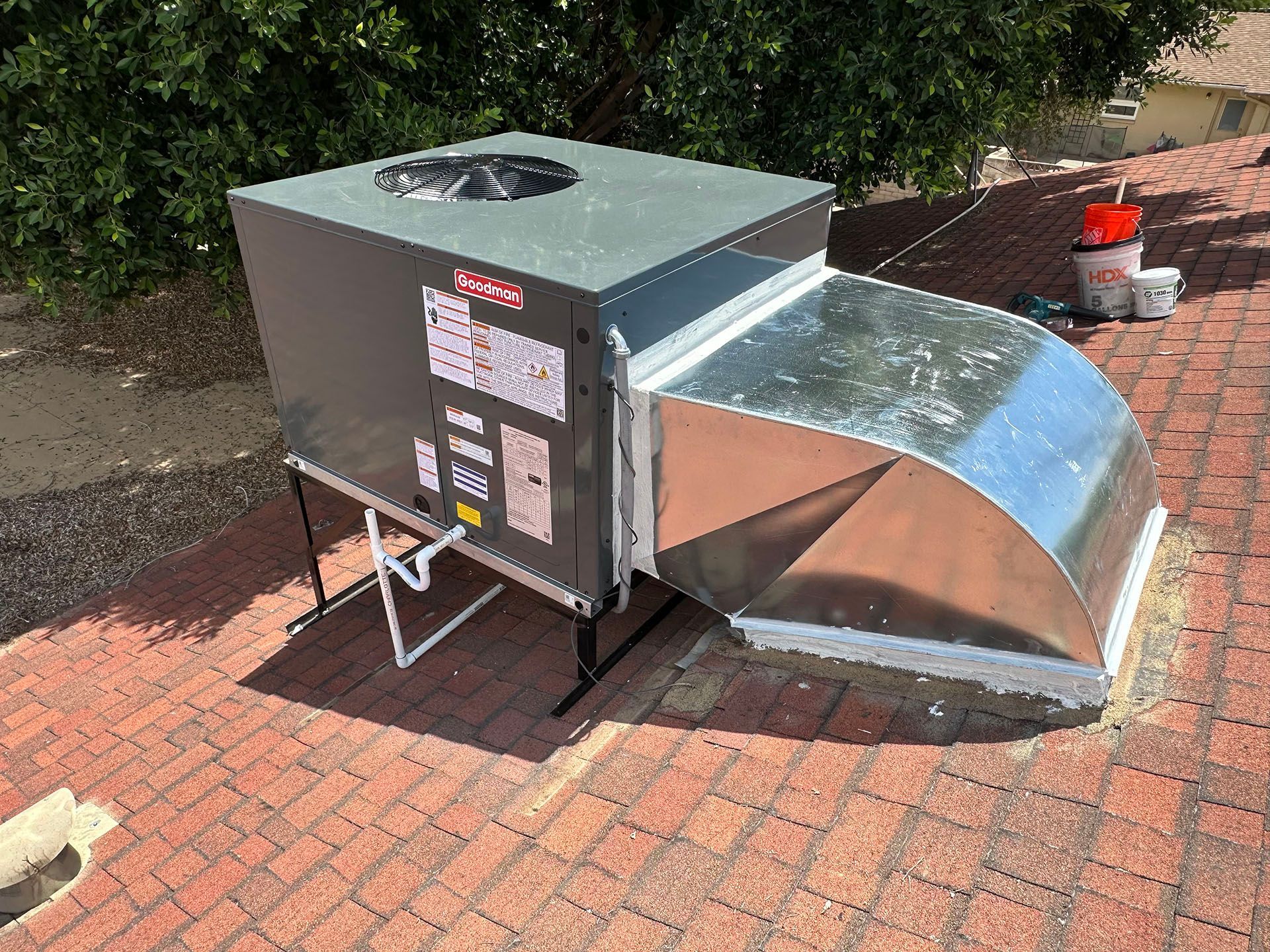 Large outdoor HVAC unit with shiny metal ducting on a brick patio, beside bushes.