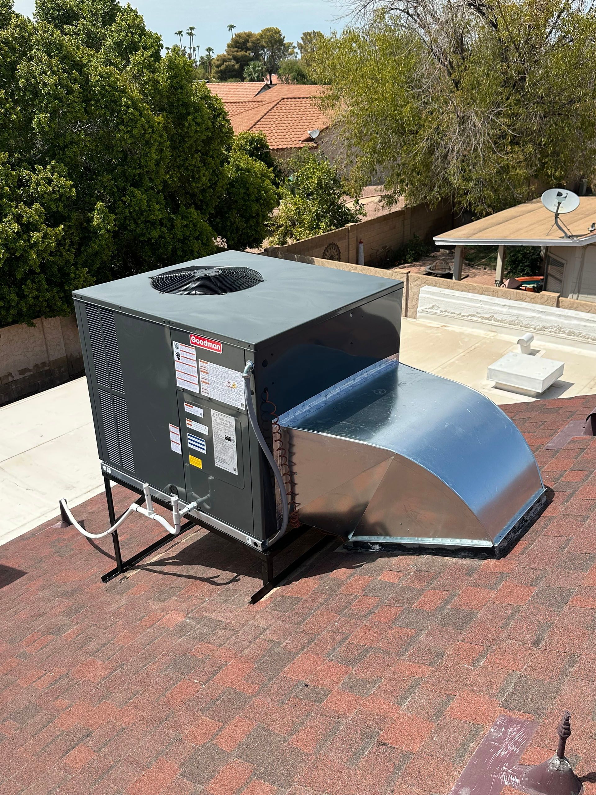 Outdoor HVAC unit on a rooftop with a metal vent duct attached, surrounded by trees and neighboring houses.