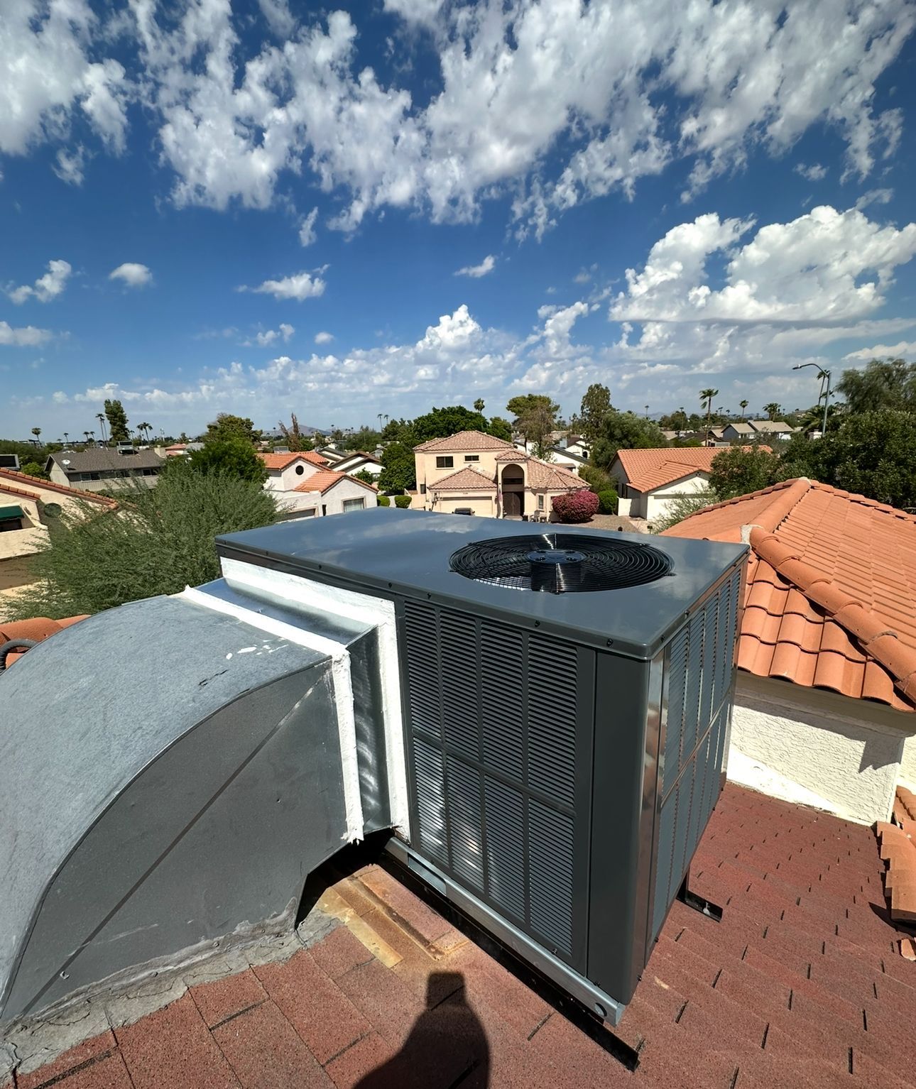 Air conditioning unit on a red tile roof, blue sky with clouds in the background, neighborhood homes visible.