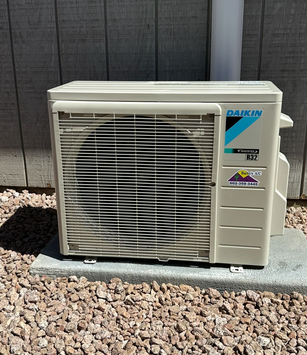 Daikin air conditioner unit, beige, mounted on gravel near a wooden wall.