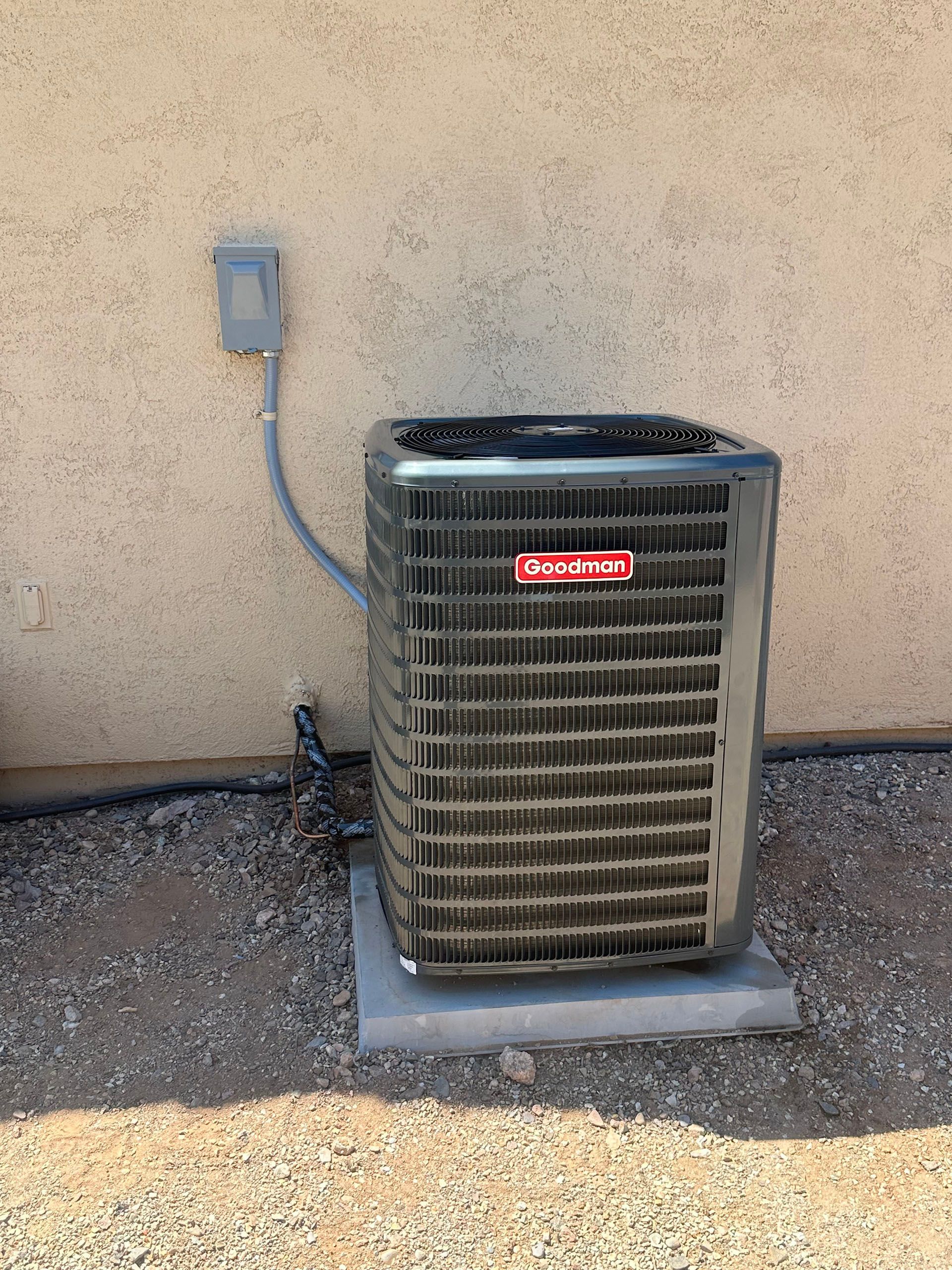 Outdoor air conditioner unit beside a stucco wall with a conduit box and gravel ground.