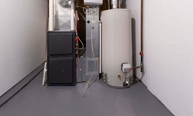 A furnace and water heater in a basement corner, gray and white, with exposed pipes and ductwork.