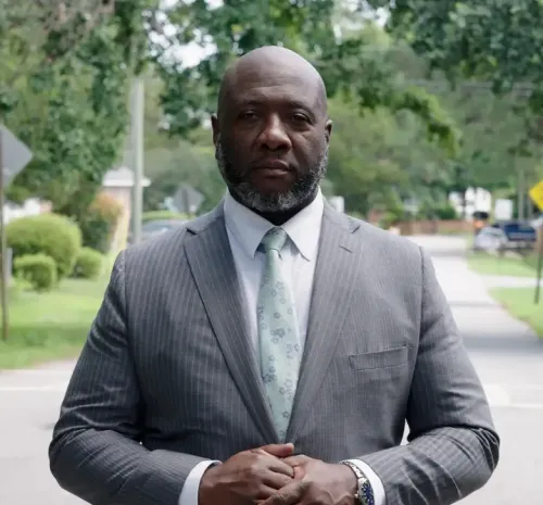 Man in a gray suit and mint tie stands on a tree-lined sidewalk, hands clasped.