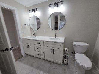 Bathroom with dual sinks, mirrors, and toilet. White vanity, black fixtures, gray walls, and light floor tile.