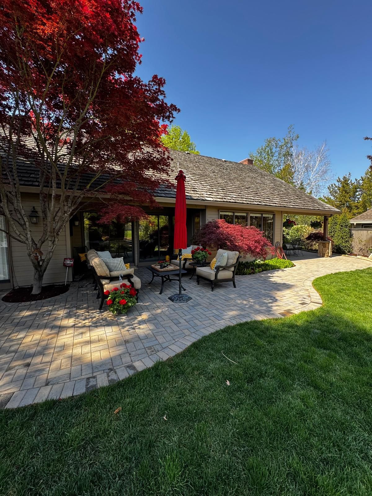 Patio with outdoor seating beside a house, red umbrella, potted flowers, and lush green lawn under blue sky