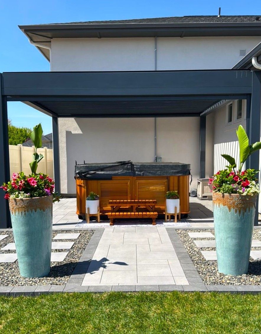 Backyard patio with a covered seating area, wooden table, and two flower-filled planters on a stone path.