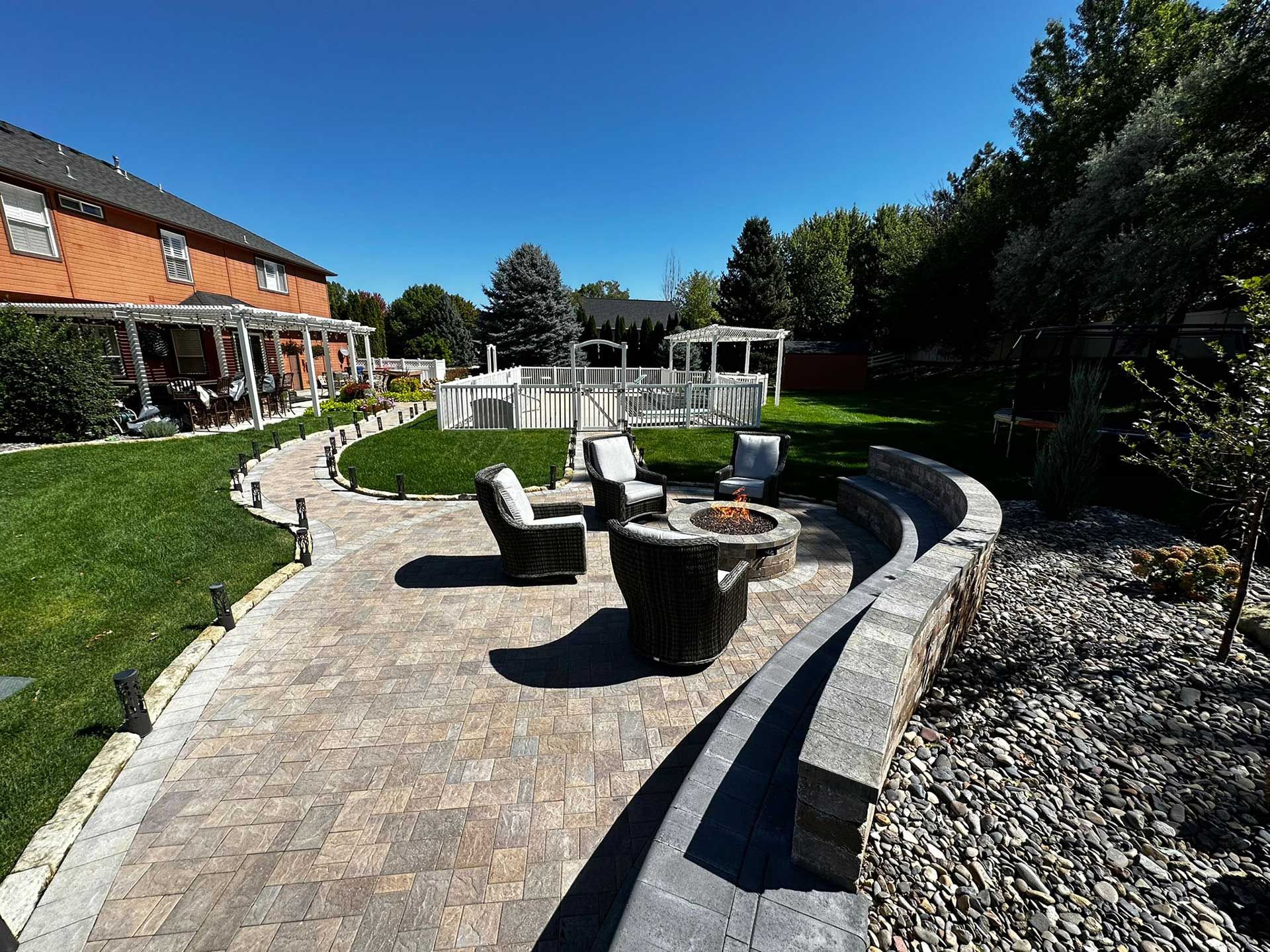 Curved stone patio with fire pit seating in a landscaped backyard beside a house.
