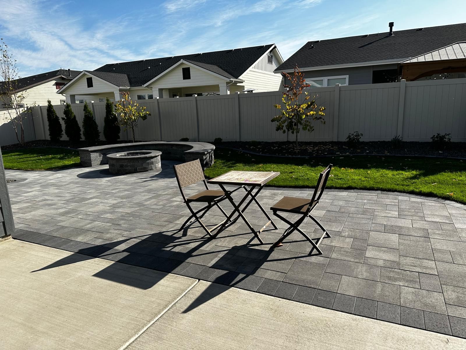 Patio with black table and chairs on a paved backyard, grass, fence, and houses in the background
