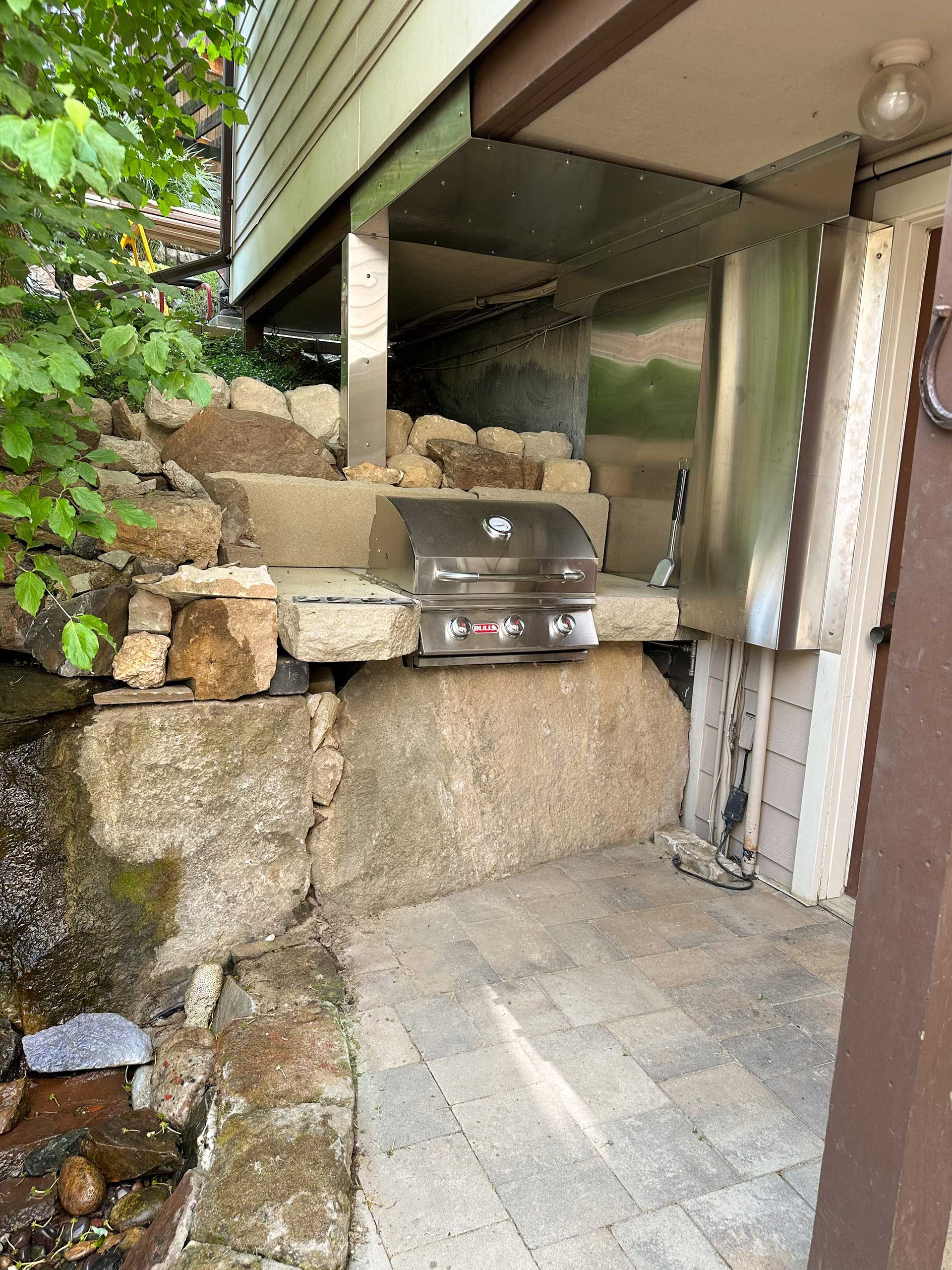 Propane grill on a stone patio under a covered deck, beside stacked rocks and greenery