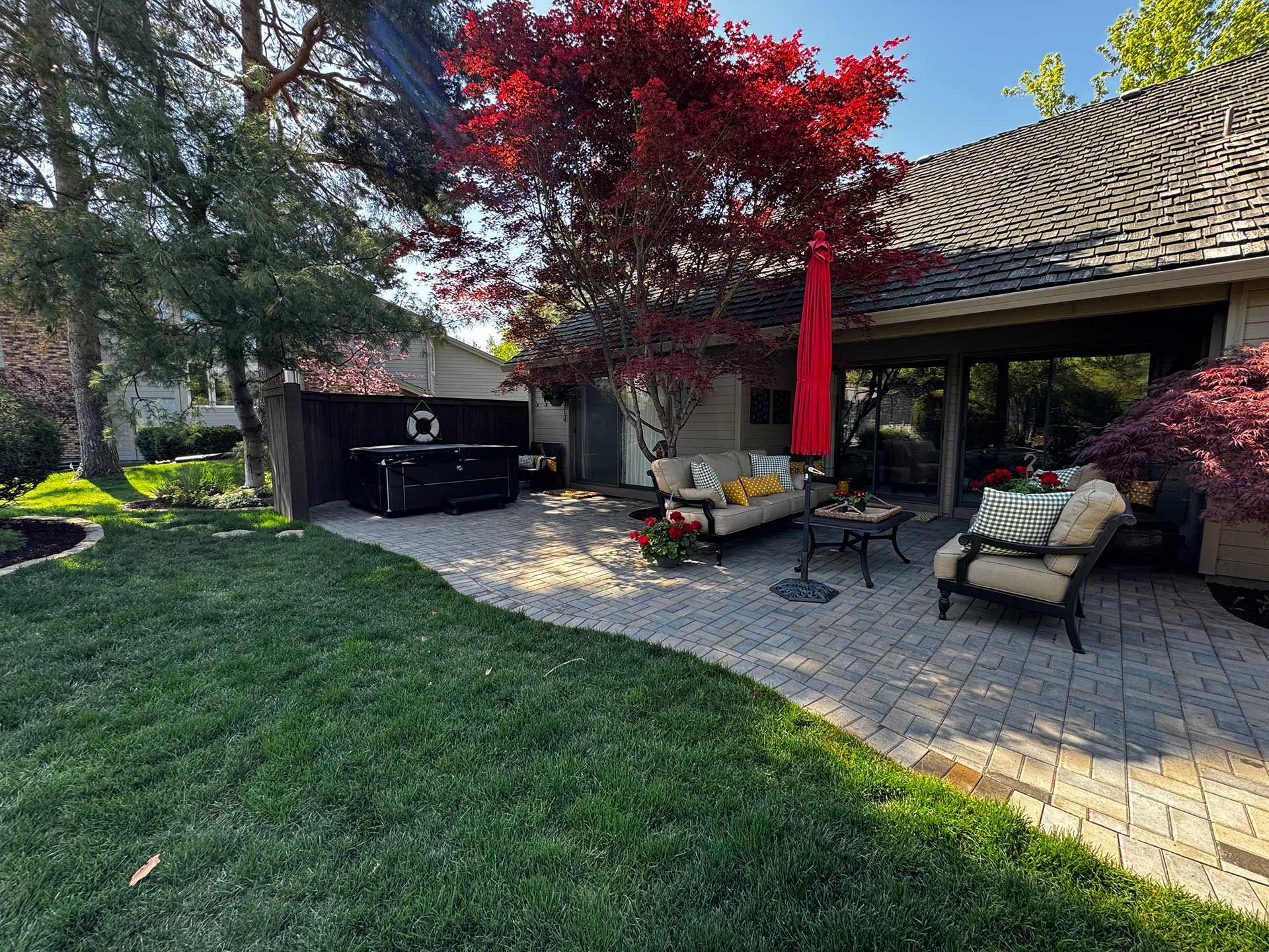 Backyard patio with lounge chairs, red umbrella, grill, and a red-leaf tree beside a stone house