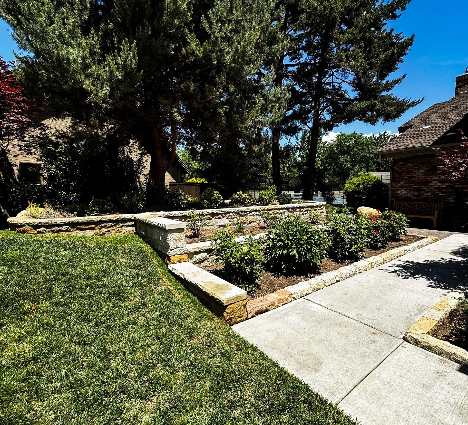 Sunny garden with stone planter beds, green lawn, trees, and a concrete walkway beside a brick house