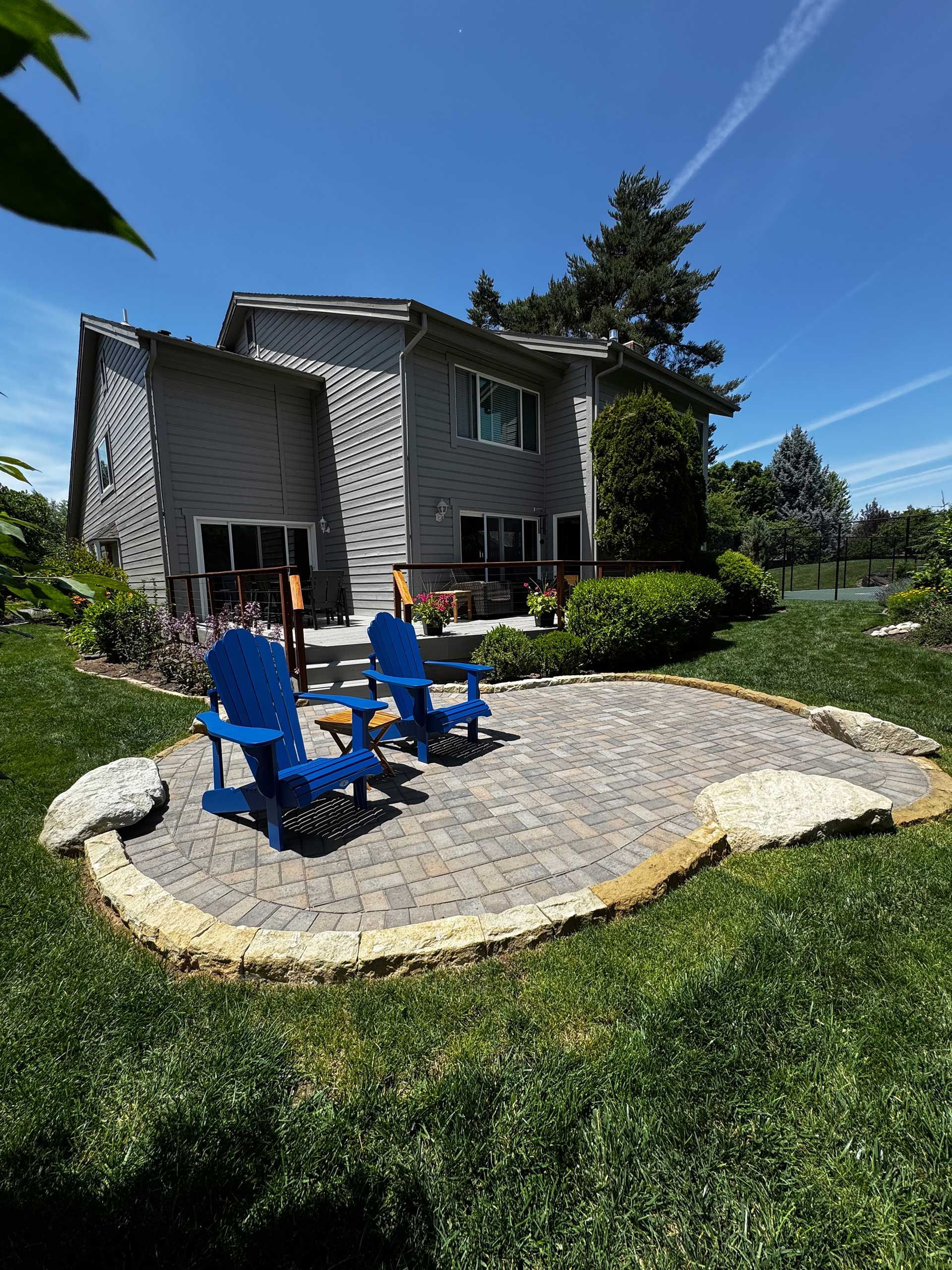 Backyard patio with two blue Adirondack chairs on a stone terrace beside a house and lawn