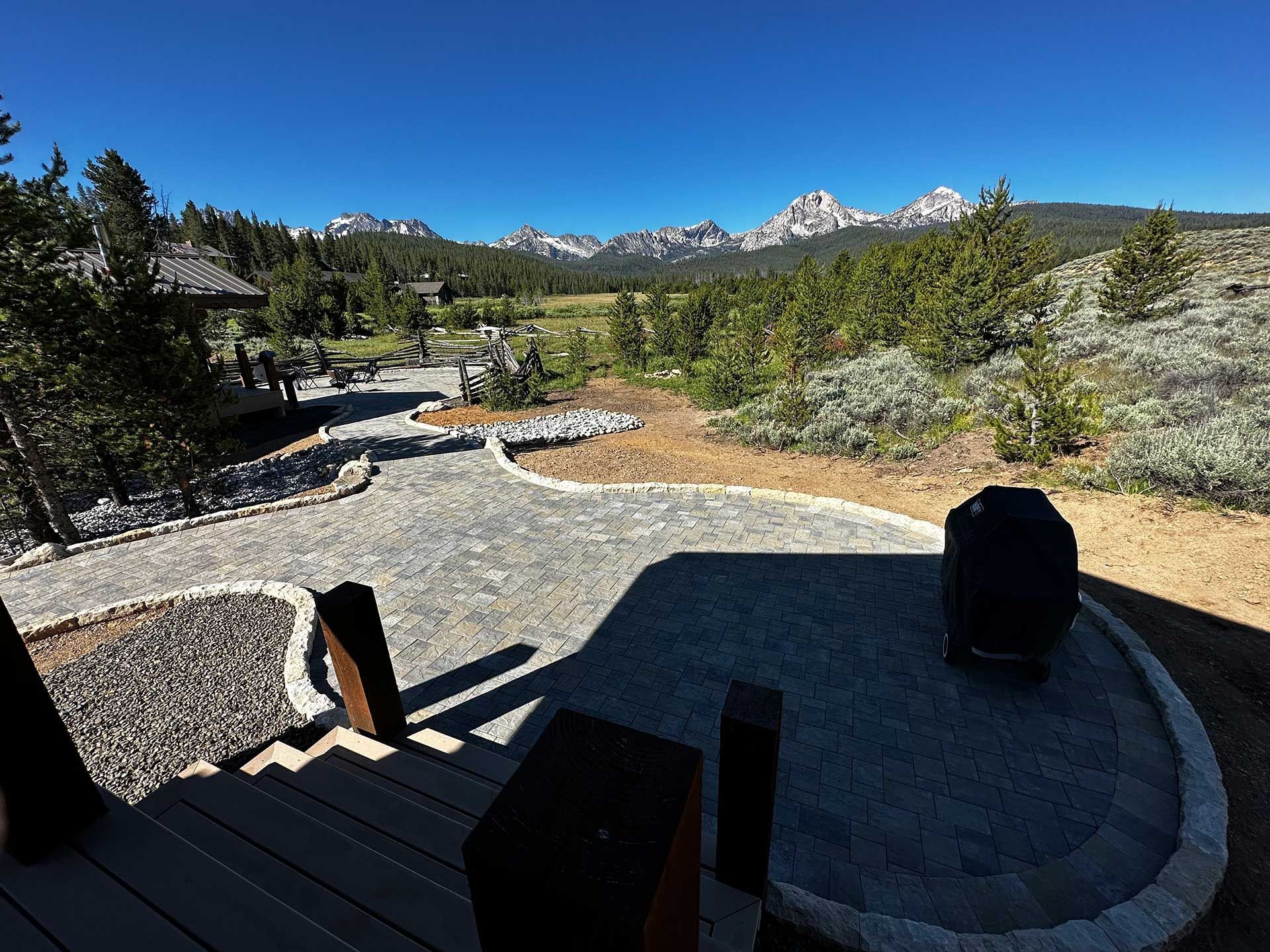 Wooden deck overlooks a gravel patio, trees, and distant snow-capped mountains under a clear sky.